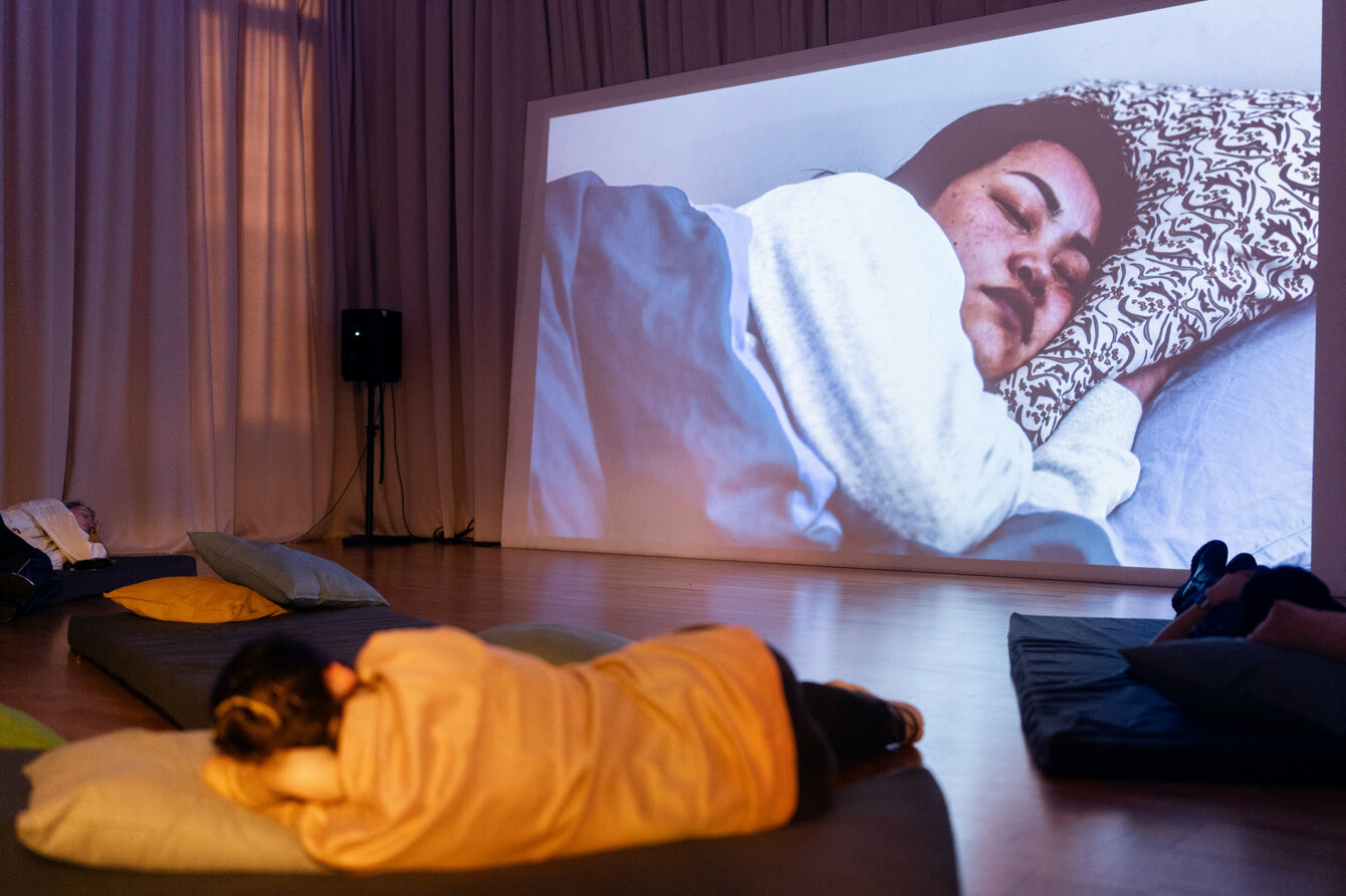 Eine große Filmprojektion fällt auf die Wand auf der zu sehen ist, wie eine Person, den kopf auf ein Kopfkissen gelegt, am schlafen ist. Die Betrachtenden der Films liegen ebenfalls in ähnlicher Position im Raum auf Kissen und  bequemen Unterlagen.