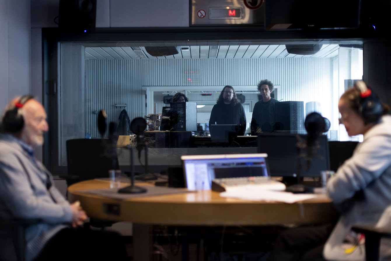 Ein älterer Herr sitzt gegenüber einer Frau in einem Tonstudio, beide tragen Kopfhörer, hinter einer Glaswand sind zwei Personen zu sehen, die die beiden beobachten.