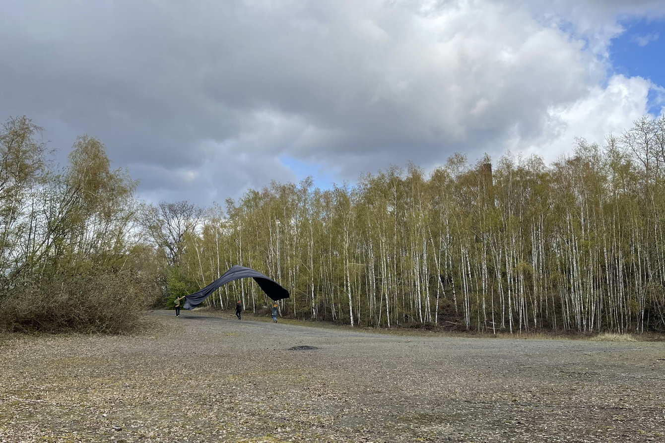 Eine Person zieht eine große schwarze Stofffahne hinter sich her, Birkenwald auf der Zeche Zollverein.