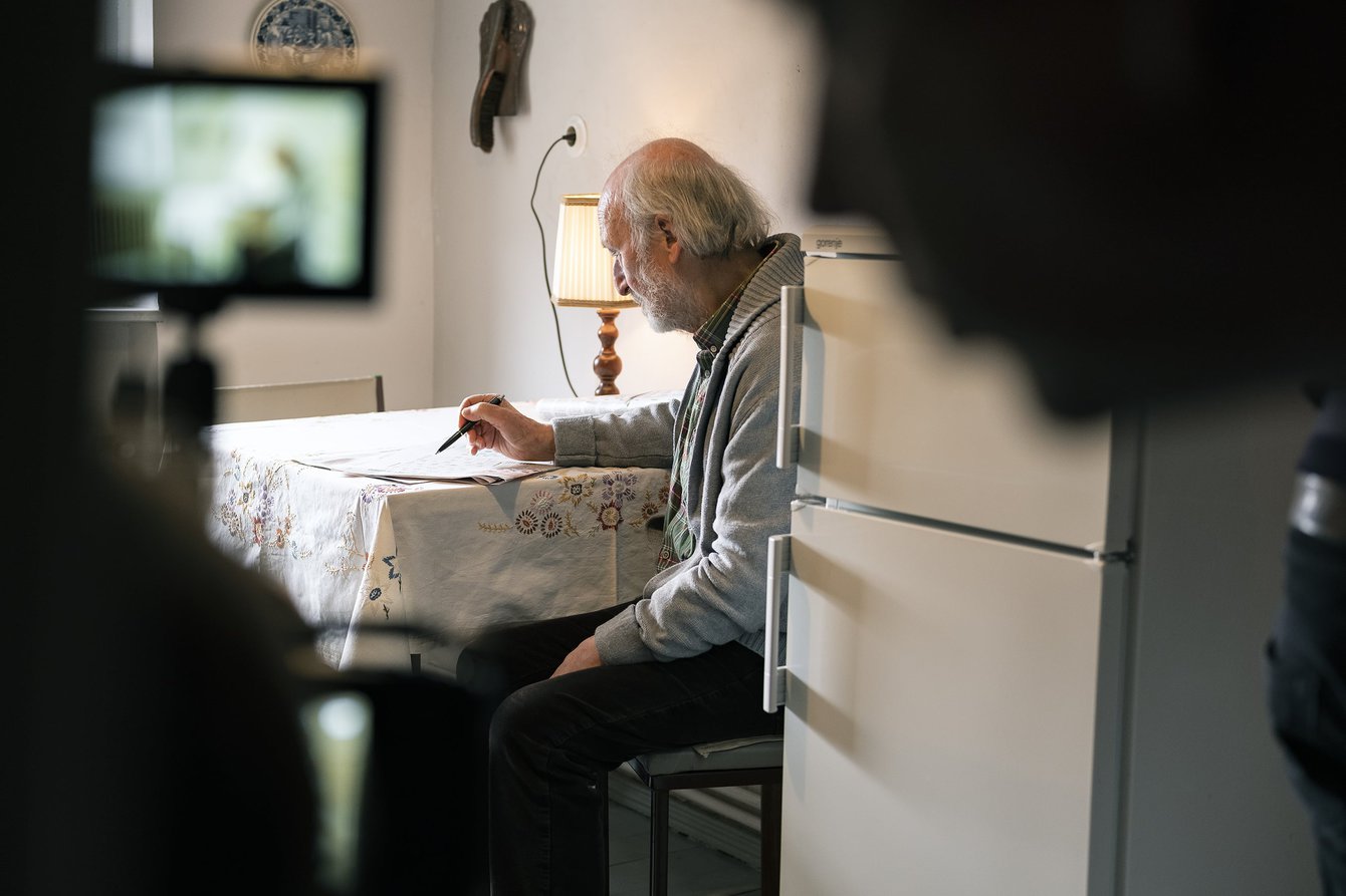 Ein älterer Herr sitzt in einer Küche und schreibt etwas auf ein Blatt Papier, im Vordergrund sind verschwommen eine Kamera und eine Person zu erkennen. 
