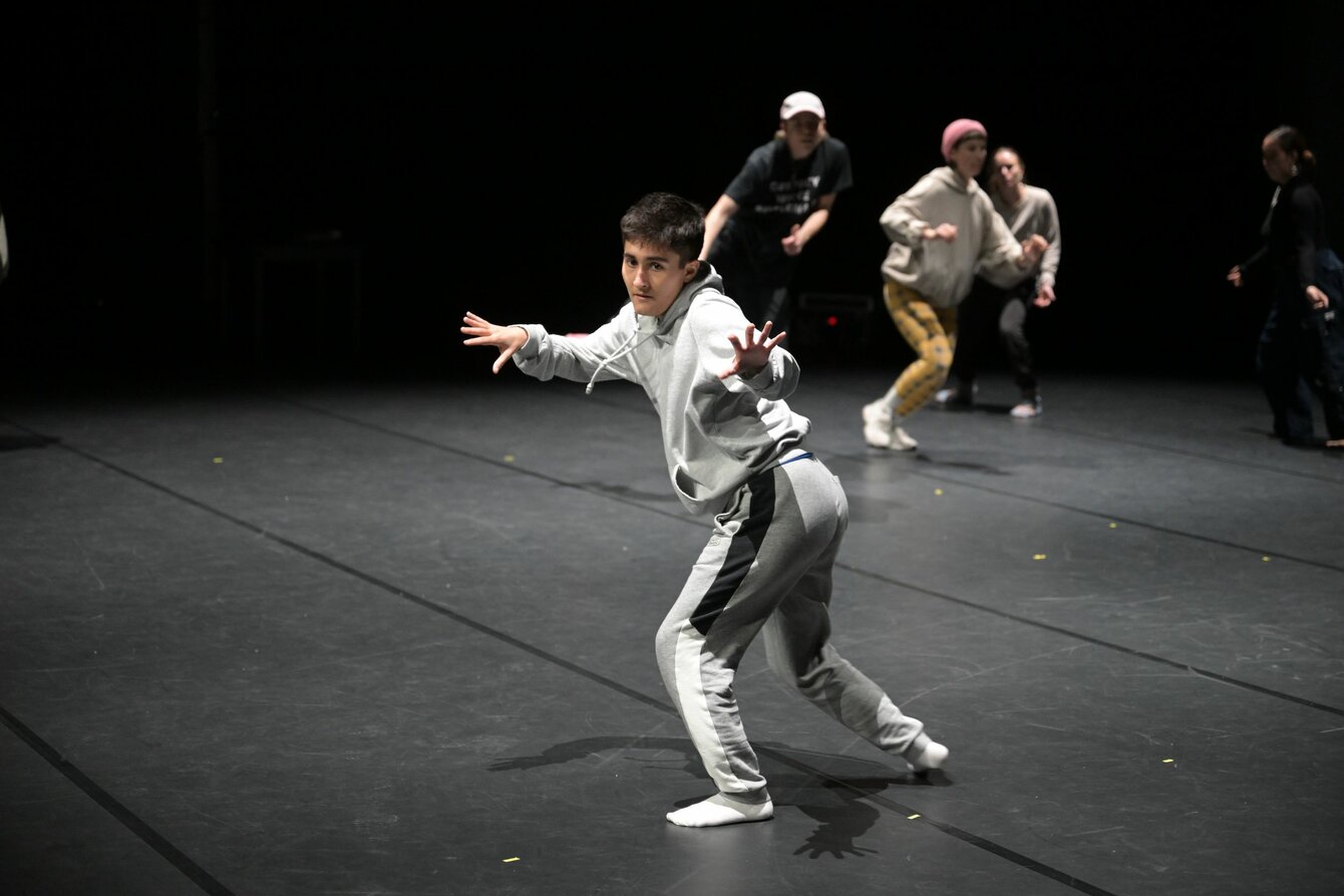 Eine Tanzaufführung auf einer Bühne. Ein Kind in einem grauen Sportanzug steht im Bildmittelpunkt seitlich in einer leichten Hocke und kleinem Ausfallschritt, die Arme und Finger leicht vom Körper gespreizt, der Blick ist fokussiert.