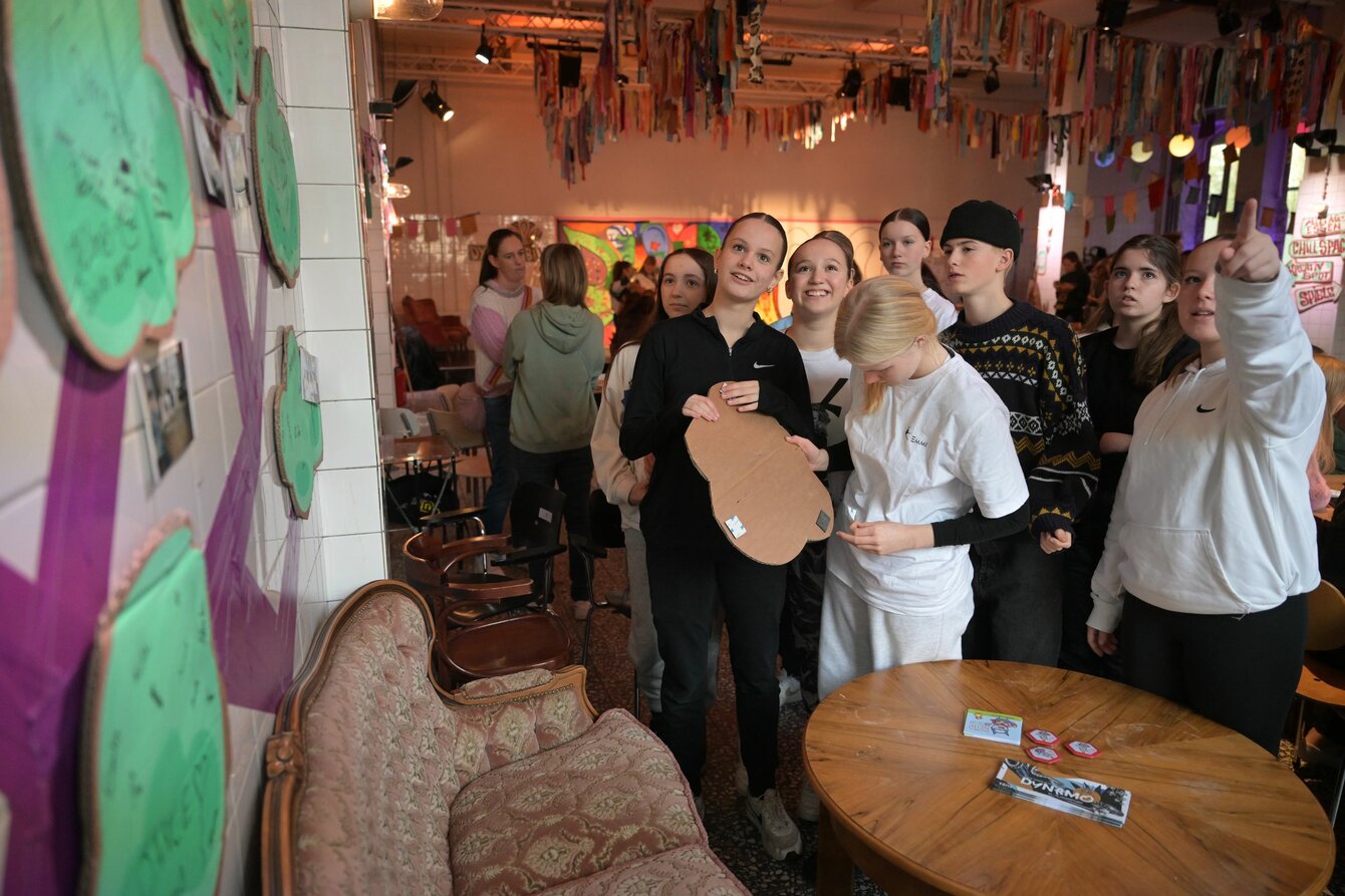 Eine Gruppe Kinder und Jugendlicher dekoriert einen Innenraum. Sie schauen und zeigen auf eine Wand, an der Schilder aufgehangen werden sollen.