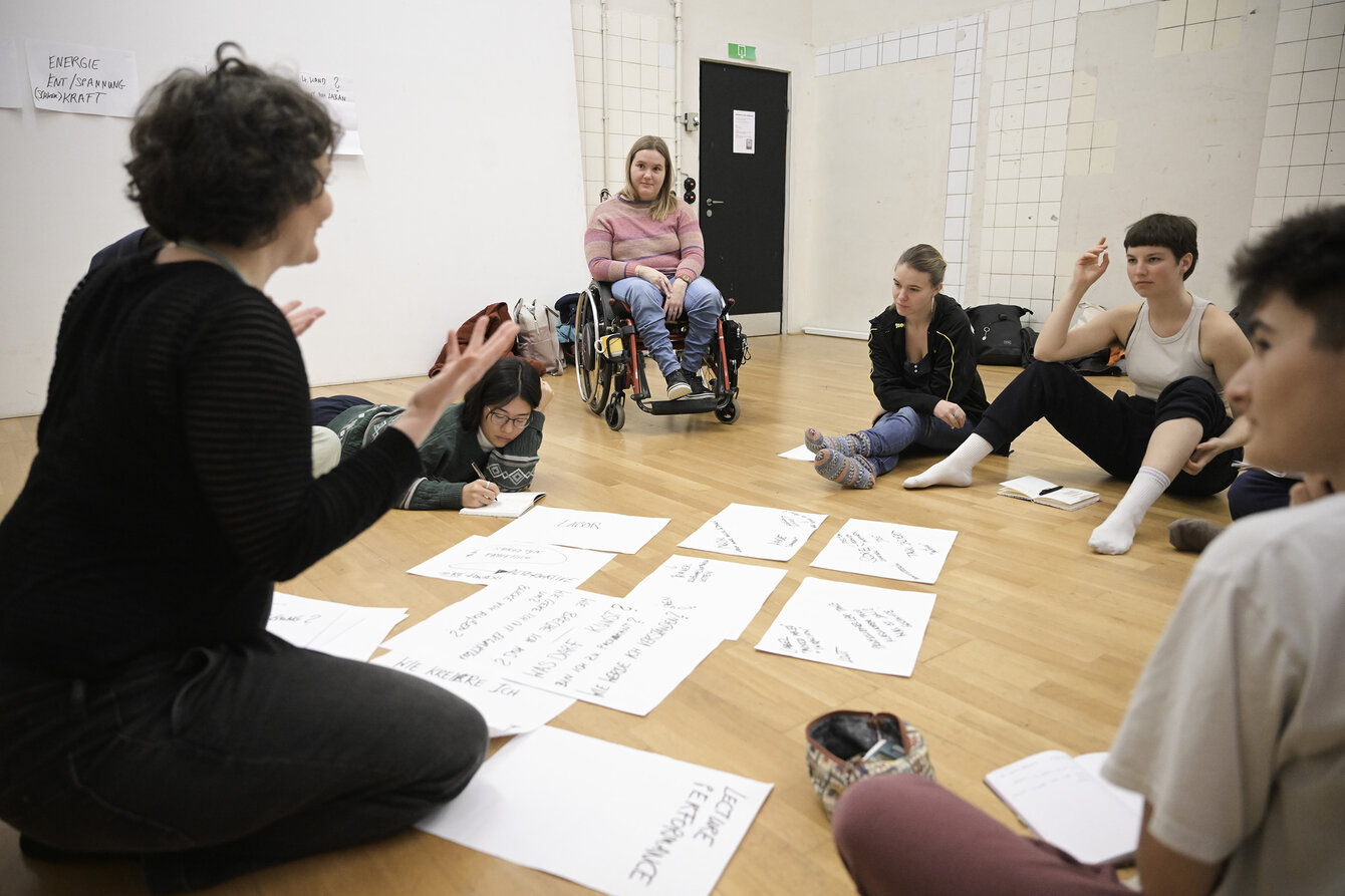 Mehrere Personen sitzen in einem Kreis zusammen und reden miteinander. In der Mitte liegen beschriebene Blätter Papier. Eine Person meldet sich.