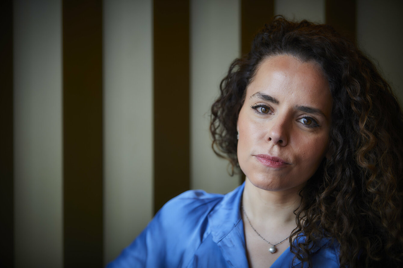 Ein Portrait von Alena Jabarine. Alena hat dunkle, lange, lockige Haare, die sie offen trägt. Sie hat ein blaues Seidenhemd an und trägt eine Kette um den Hals. Hinter ihr eine creme-braun gestreifte Wand.