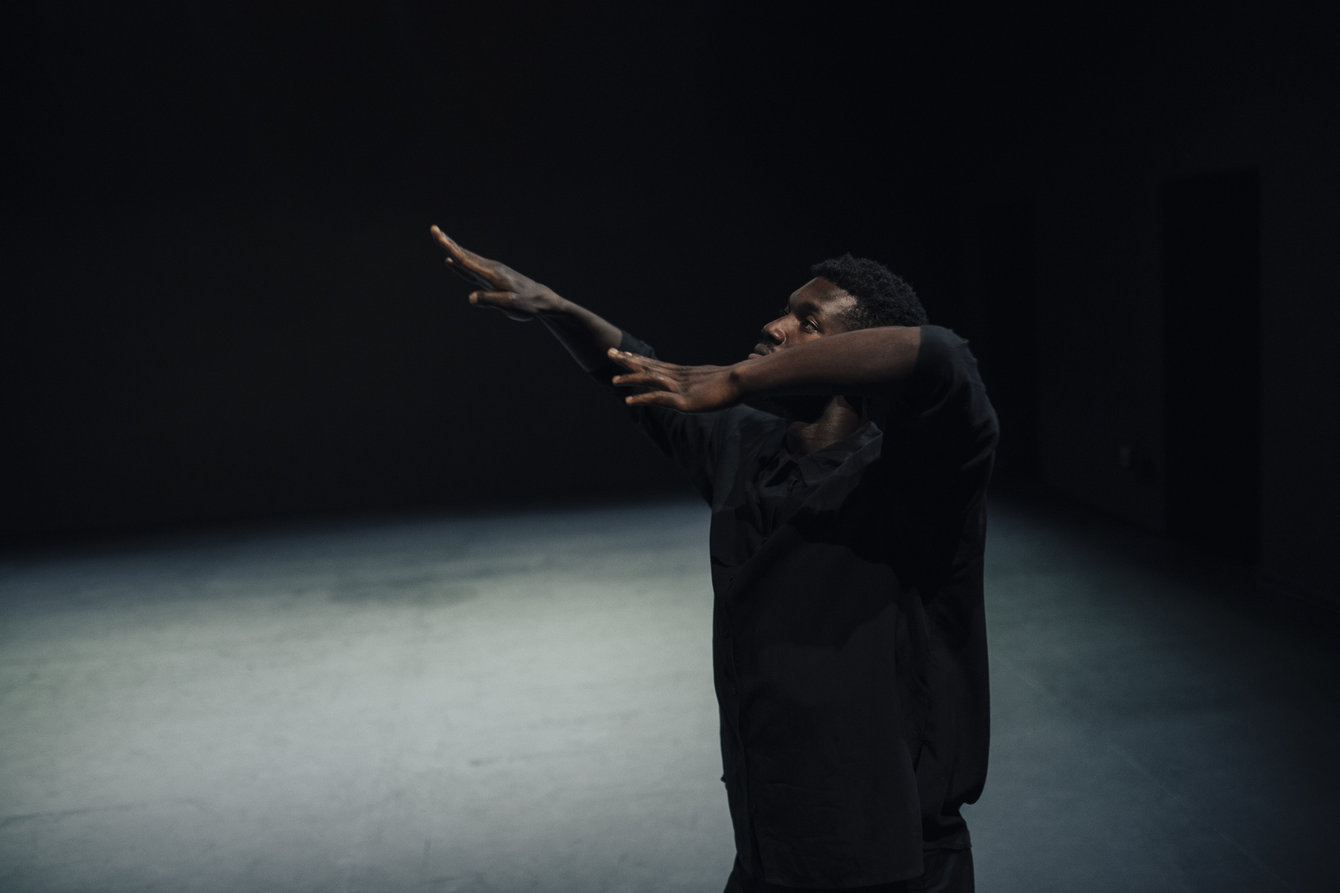 Ein Tänzer mit Armbewegungen vor schwarzem Hintergrund in schwarz gekleidet