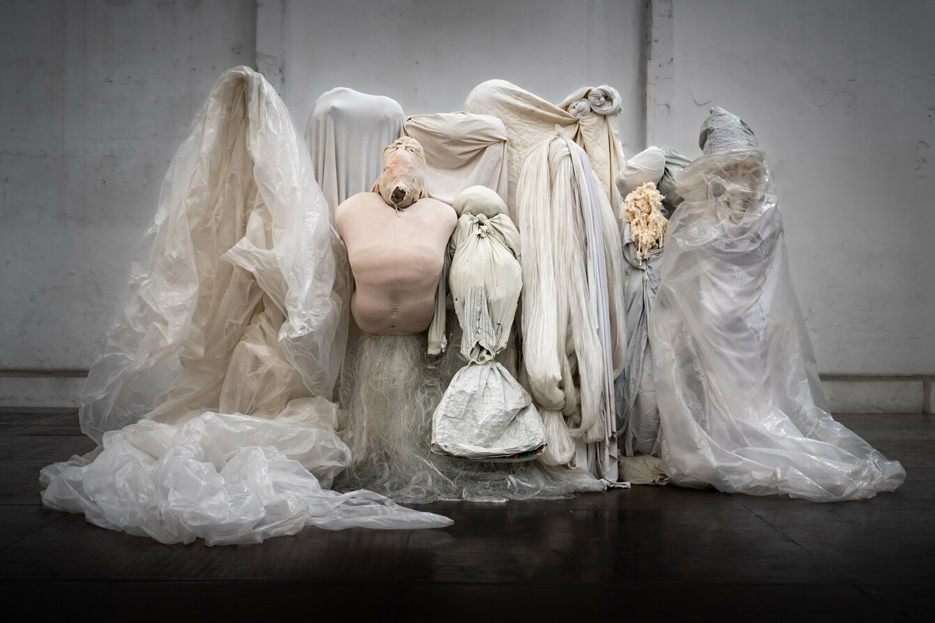 Mehrere verhüllte Figuren aus Stoff, Plastik und anderen Materialien stehen dicht beieinander. Einige wirken wie geisterhafte Gestalten, andere haben verzerrte menschliche Formen.