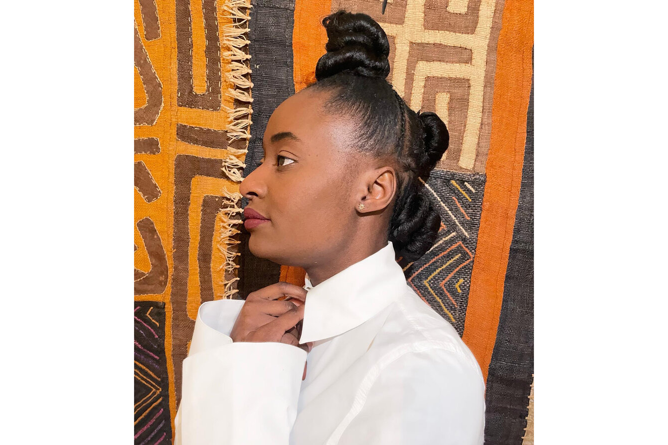 Ein Portrait von Fifi Mvunuku. Es zeigt Fifi von der Seite. Fifi trägt ein weißes Hemd, hinter ihr ist ein Kente-stoff zu sehen. 
