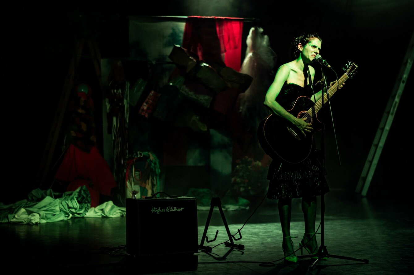 Eine weiblich gelesene Person steht mit einer Gitarre auf der Bühne, das Licht ist dunkel, im Hintergrund sind die Umrisse von Gegenständen zu erkennen