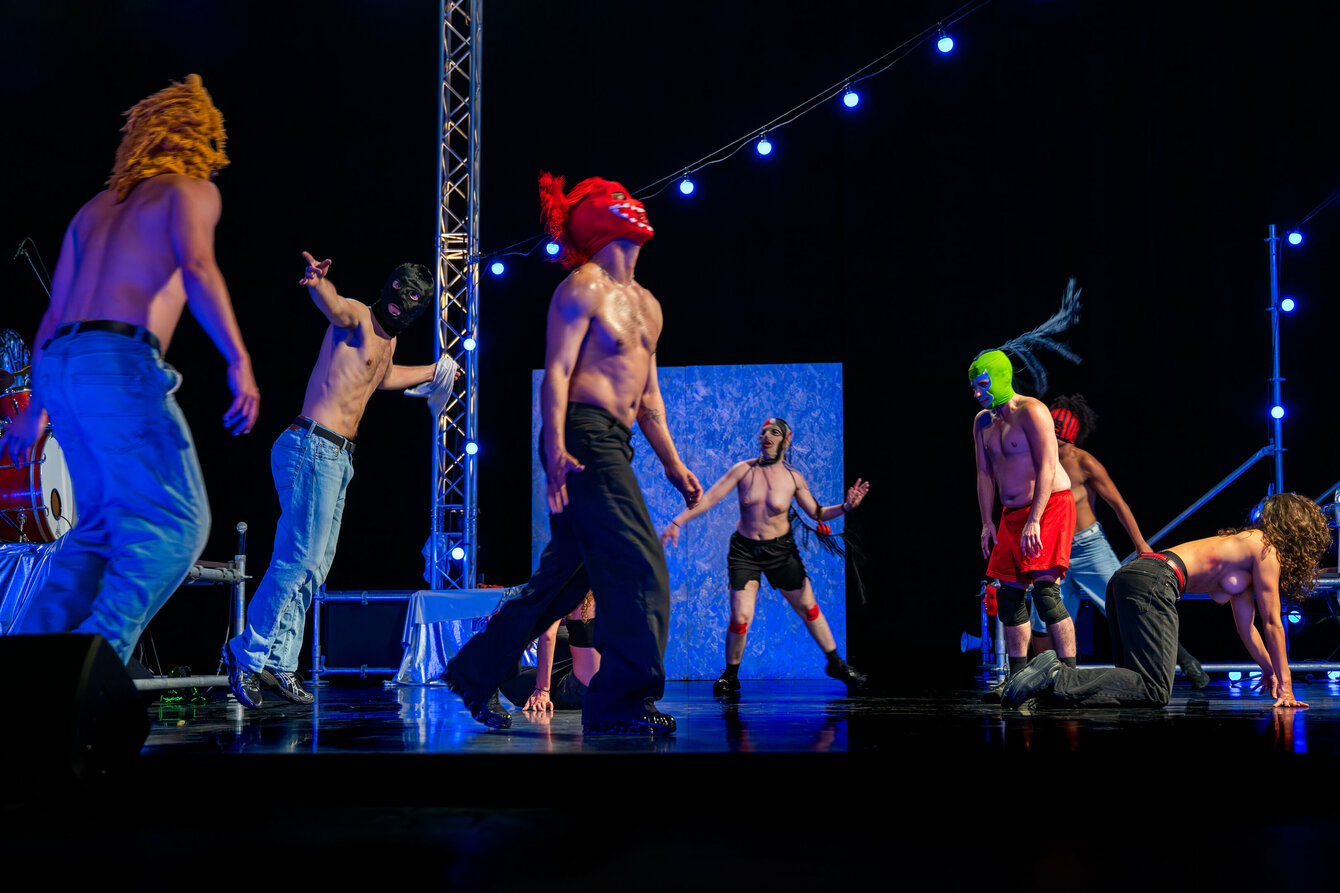 Eine Szene aus Delirious Night: Mehrere maskierte Performer*innen, oberkörperfrei, auf der Bühne. Die Bühne ist teils in blaues Licht getaucht. Sie tanzen ekstatisch.