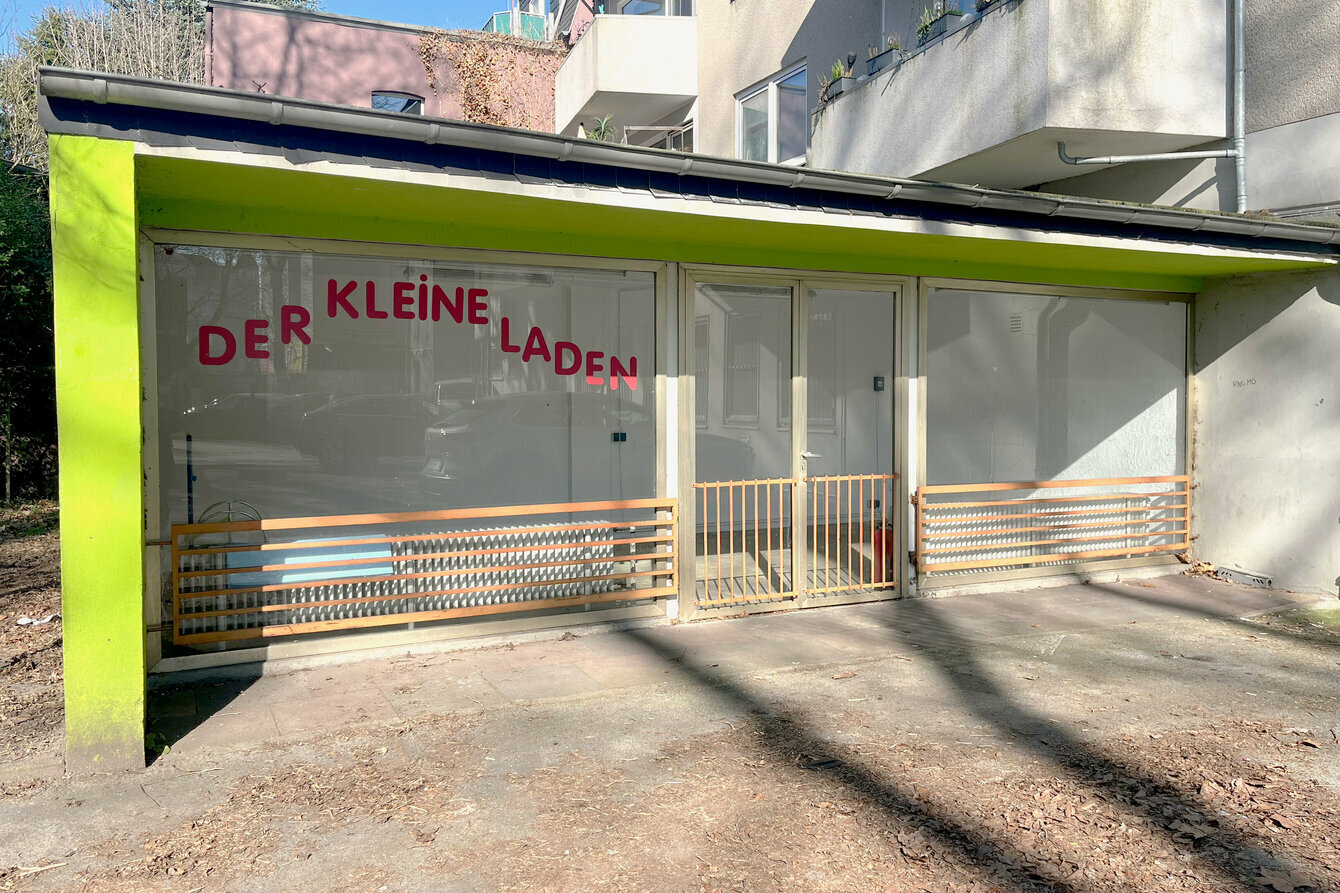 Fotografie des Kleinen Ladens. In einem Hinterhof ist der Ausstellungsraum frontal abgebildet, der längliche Ausstellungsraum ist von dort aus betrachtet komplett verglast und wärt einen Blick ins Innere. Auf der Scheibe ist der rote Schriftzug ›Der Kleine Laden‹ angebracht. Die sichtbare Außenwand auf der linken Seite ist grün gestrichen. 