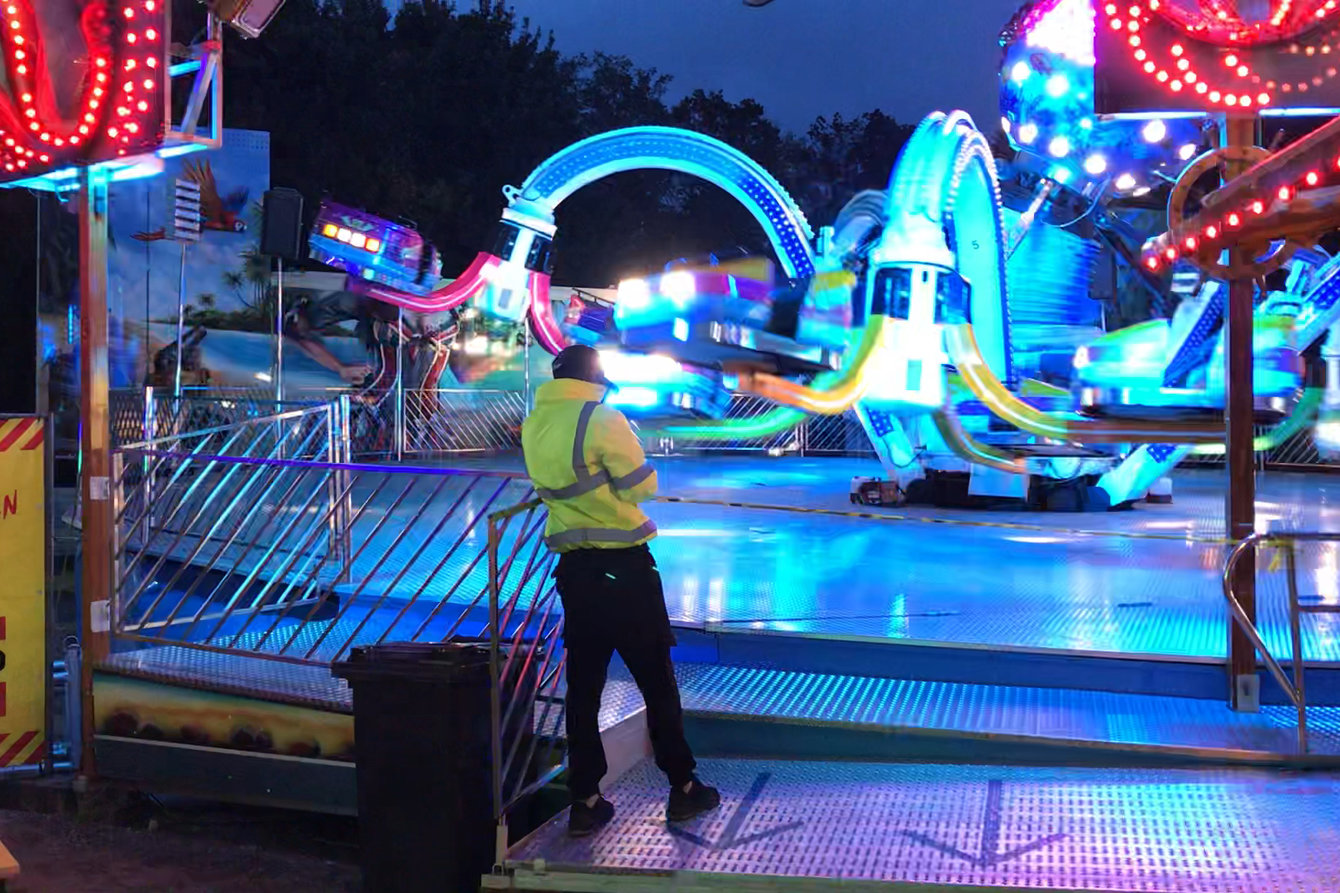 Eine dienstleistende Person trägt eine neongelbe Arbeitsjacke. Sie steht, gelehnt an ein Geländer und mit dem Rücken zur Kamera, vor einem bunt beleuchteten Fahrgeschäft.