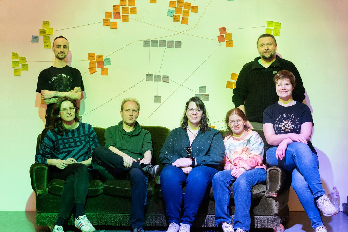 Gruppfenfoto der Projektteilnehmer:innen von I Am Your Body, sie sitzen auf einem Sofa vor einer weißen Wand mit Klebezetteln