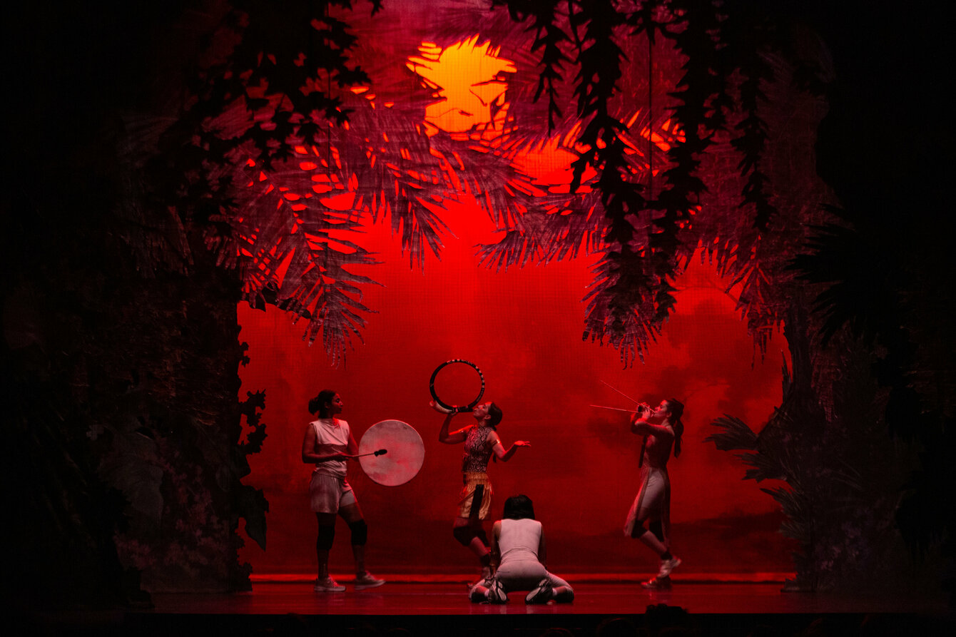 Mehrere Personen auf einer Bühne mit roter Beleuchtung und Dschungelpflanzen