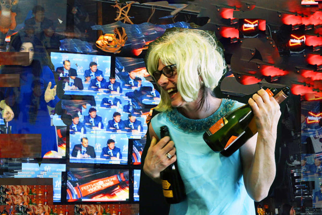 Eine Person im blauen Kleid mit Perücke und zwei Flaschen in der Hand. Die Person trägt eine blonde Perücke und eine Sonnenbrille und lacht. Hinter der Person sind verschiedene Screens zu sehen.