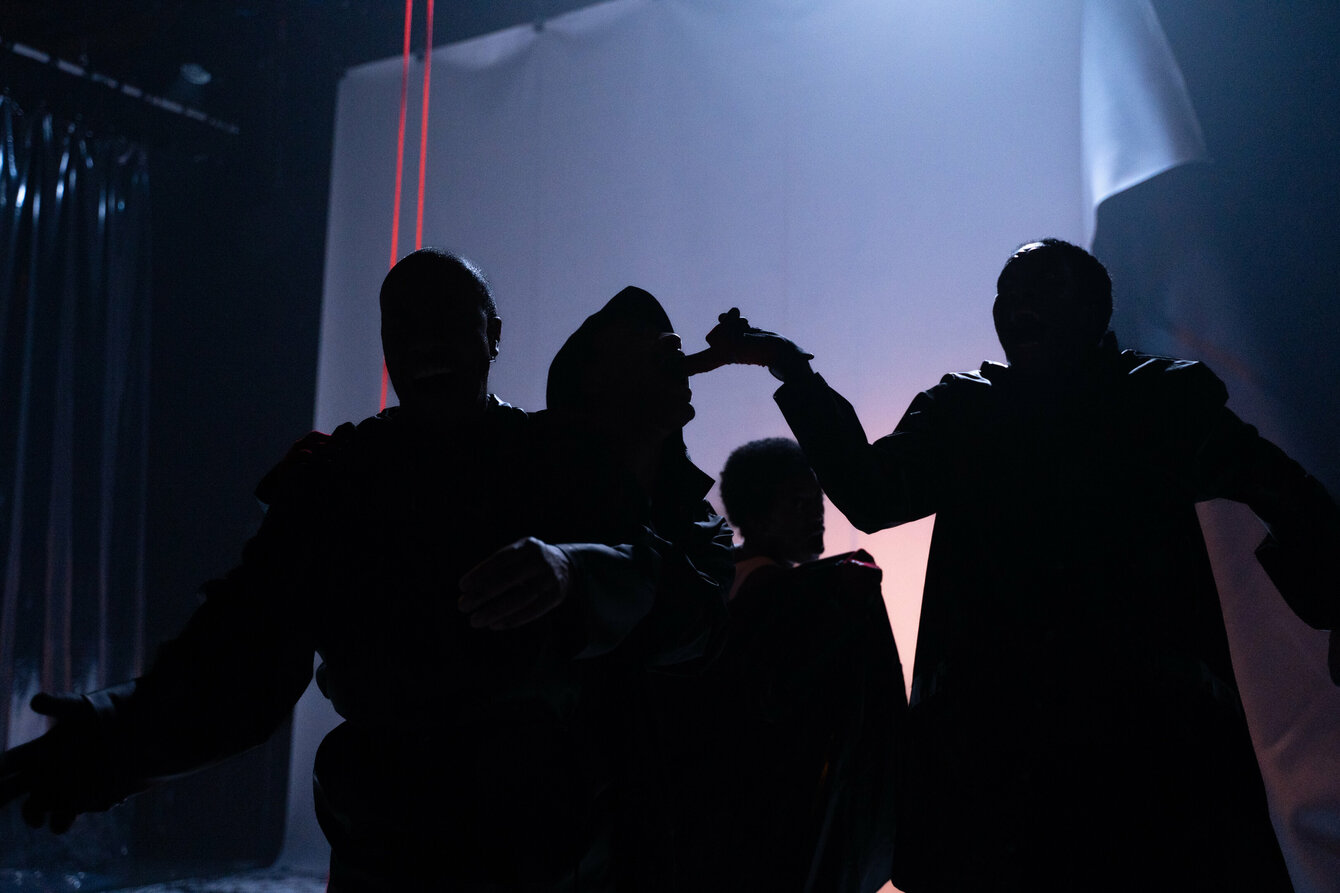 Dunkle Silhouetten einer Gruppe von Performer:innen vor einem beleuchtetem Vorhang. Die Stimmung ist düster, einige Performer:innen tragen Gewänder mit tiefen Kapuzen. Im Hintergrund sticht eine rote Schnur in der ansonsten bläulichen Tönung des Bildes hervor. 