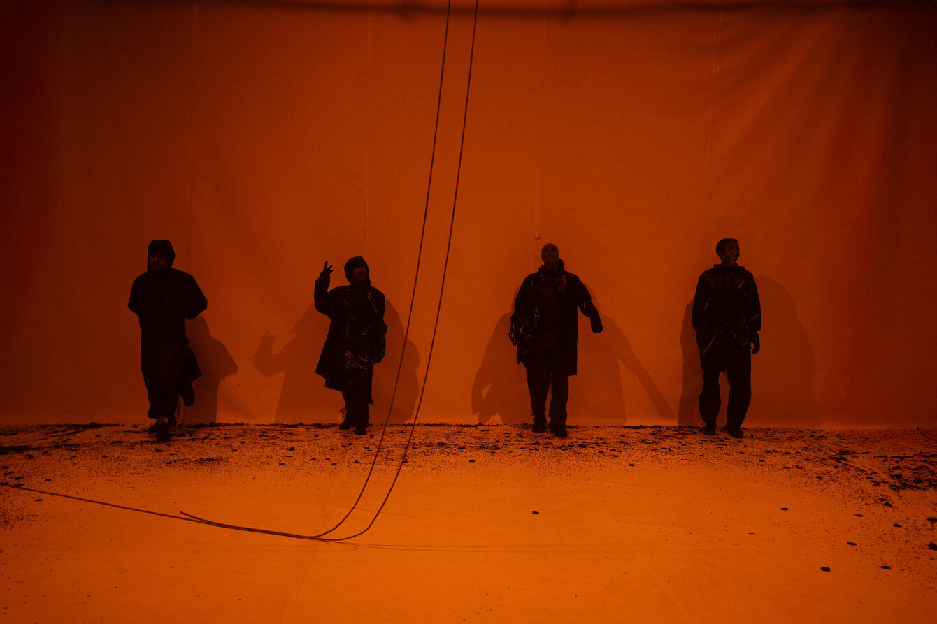 4 Performer:innen stehen in einem schwach rötlich ausgeleuchtetem Bühnenraum an der Wand. Von oben hängen mehrere Kabel herab auf den Boden.