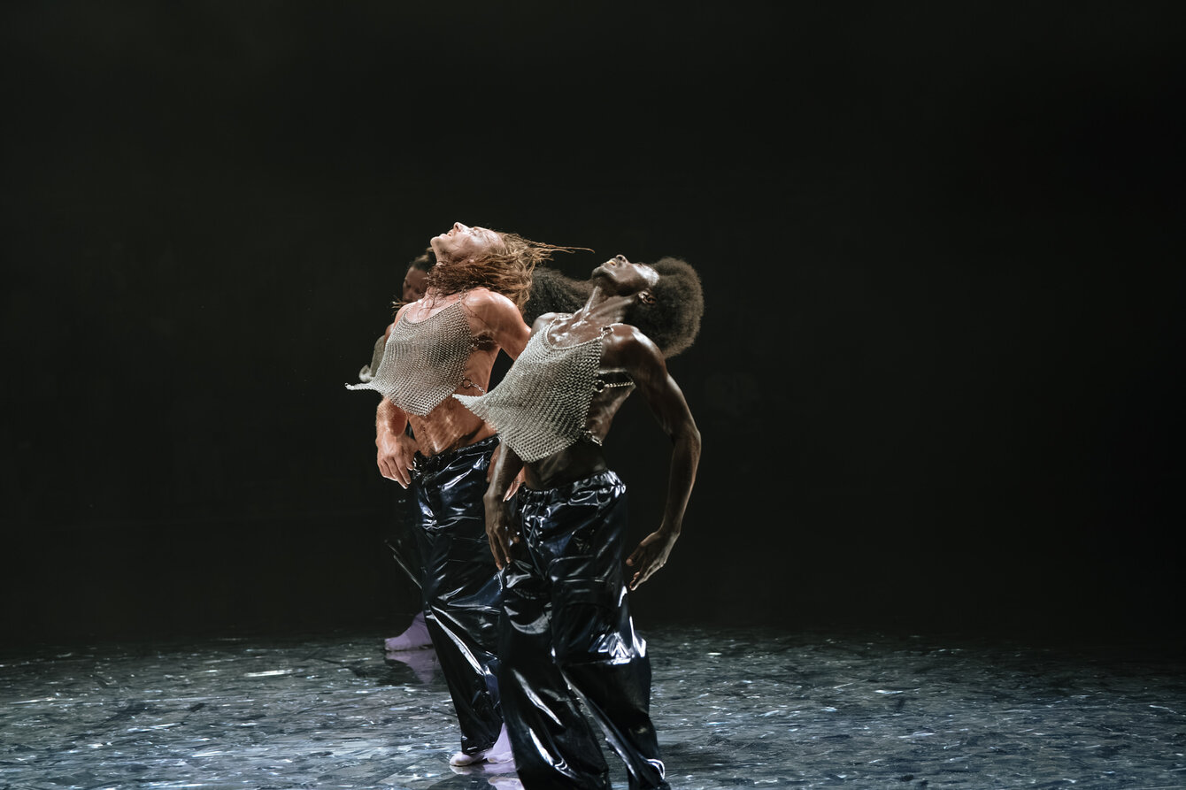 Eine Szene aus F*cking Future: Zwei Performer*innen sind beim Tanzen auf der Bühne zu sehen. Sie werfen beide die Köpfe nach hinten und strecken dabei jeweils den Brustkorb nach vorne. Beide tragen silber glitzernde, ärmellose Tops und schwarze Lackhosen.