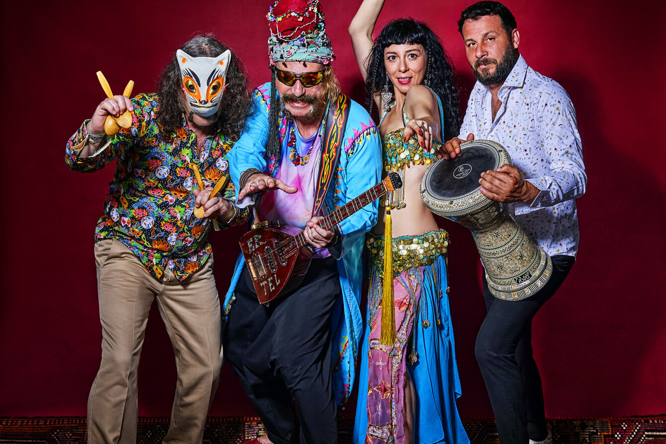 Ein Gruppenfoto der Band Baba ZuLa: Vier bunt gekleidete Personen posieren stehend vor einer roten Wand. Links eine maskierte Person mit schulterlangen Haaren und dem Musikinstrument Löffel in der Hand, daneben Murat Ertel mit dem Instrument Electric Saz, daneben eine tanzende Esma Ertel und rechts neben ihr Perkussionist Ümit Adakale.