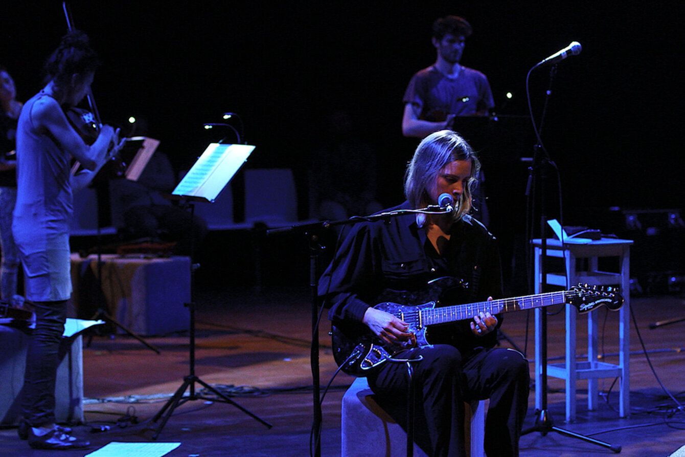 Eine bläulich erleuchtete Bühne mit mehreren Musiker:innen und einer Person mit Gitarre im Vordergrund