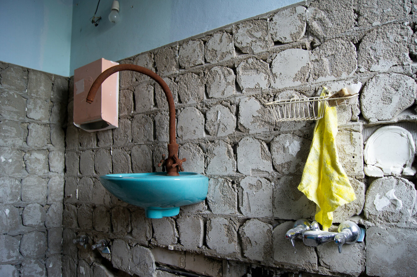 Installation von Kira Fröse: Ein Raum mit unverputzter Steinwand und hellblauer Tapete. An der Wand ein Waschbecken in Türkis mit braunem Wasserhahn. Rechts daneben hängt ein gelbes Handtuch, links neben dem Waschbecken ein Papierspender in Apricot.
