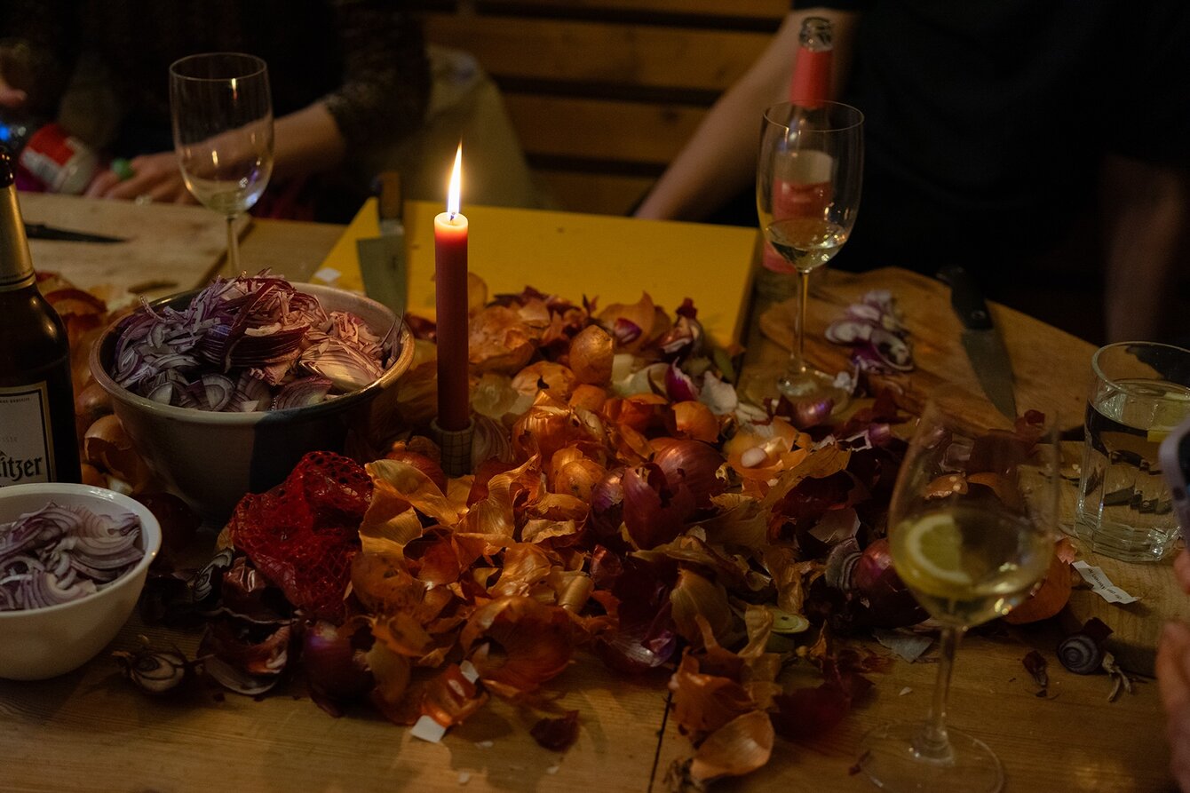 Ein gedeckter Tisch: In der Mitte eine brennende Kerze, drumherum viele Zwiebelschalen. In der linken Bildhälfte zwei Schalen mit Zwiebelringen. 