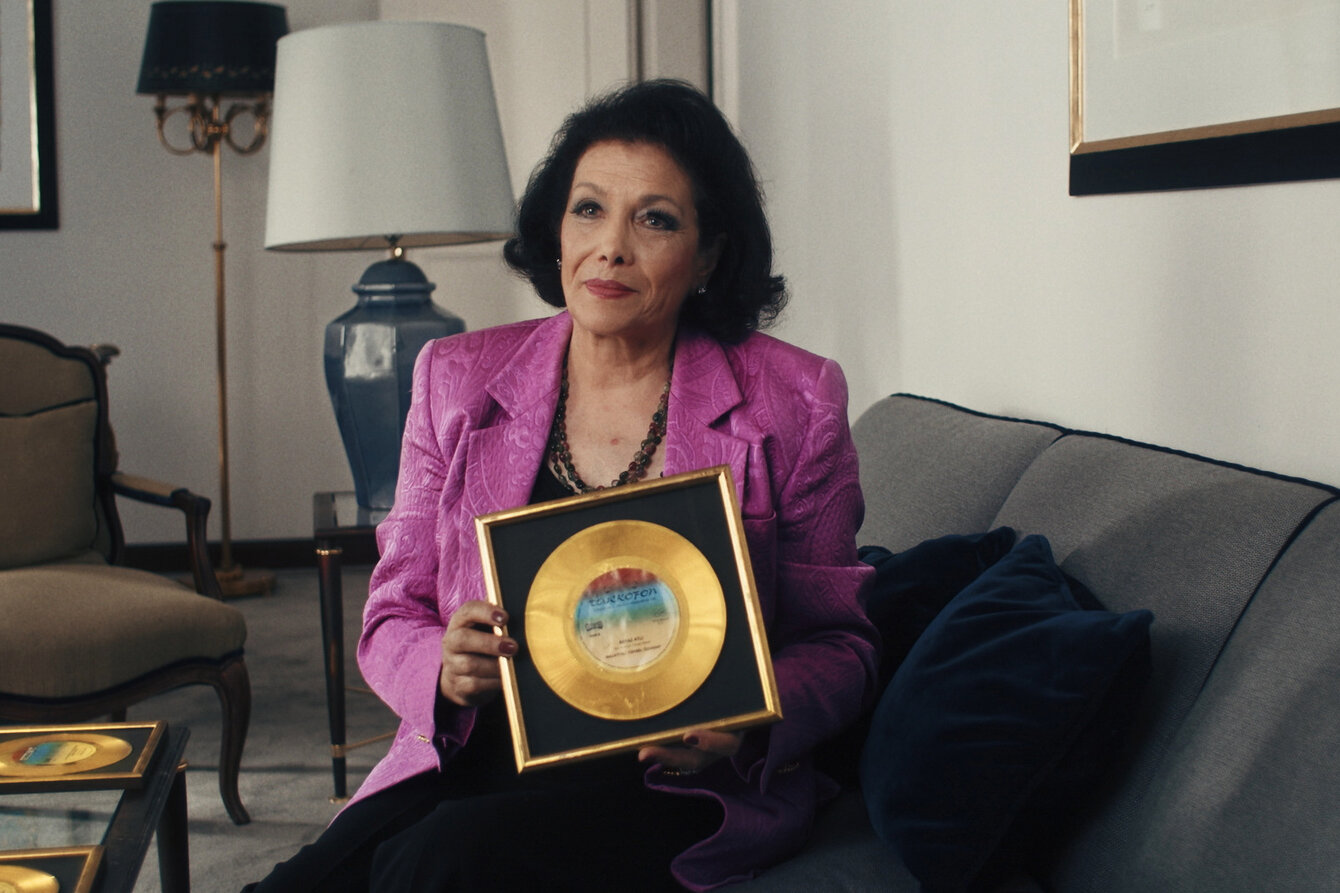 Eine Frau mit Lila Blazer und einer goldenen Schallplatte in der Hand sitz auf einem Sofa