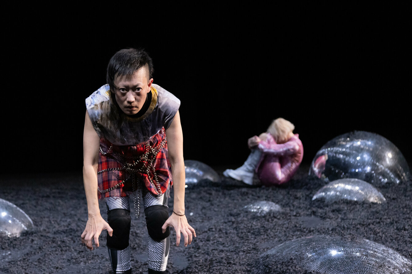 Eine Szene aus der Performance Glitch Witch. Das Bild zeigt zwei Performer*innen auf einer dunklen Bühne. Links im Vordergrund ein*e Performer*in, die*der sich nach vorne bückt und in die Kamera blickt. Im Hintergrund eine Person, die mit angezogenen Beinen und zwischen den Armen vergrabenem Kopf auf dem Boden kauert. Diese Person ist von der Seite zu sehen. 