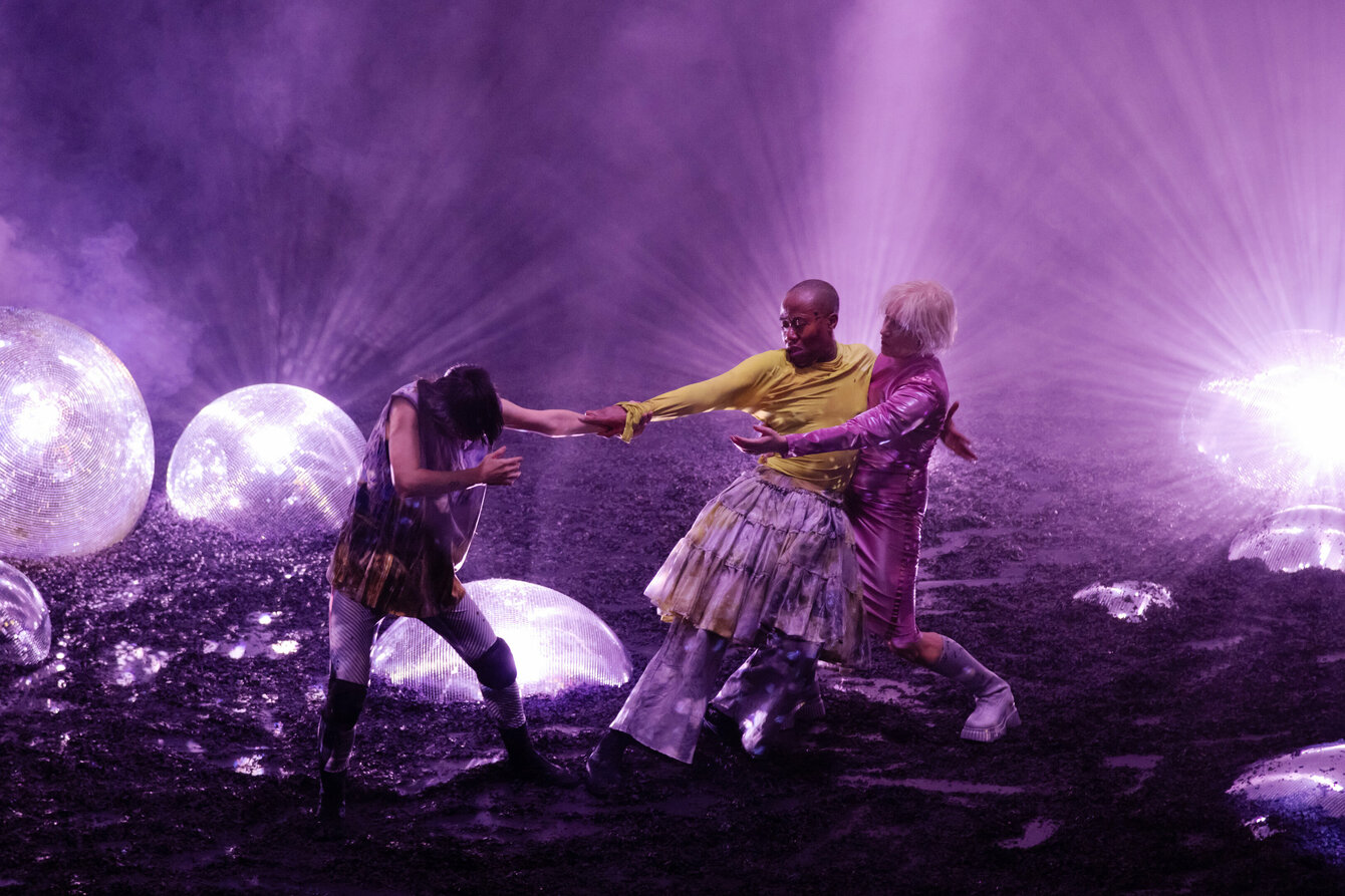 Eine Szene aus der Performance GLITCH WITCH. In der Bildmitte drei Performer*innen auf einer Bühne, die in lila Licht getaucht ist. Auf dem Bühnenboden sind riesige, leuchtende Diskokugeln zu sehen