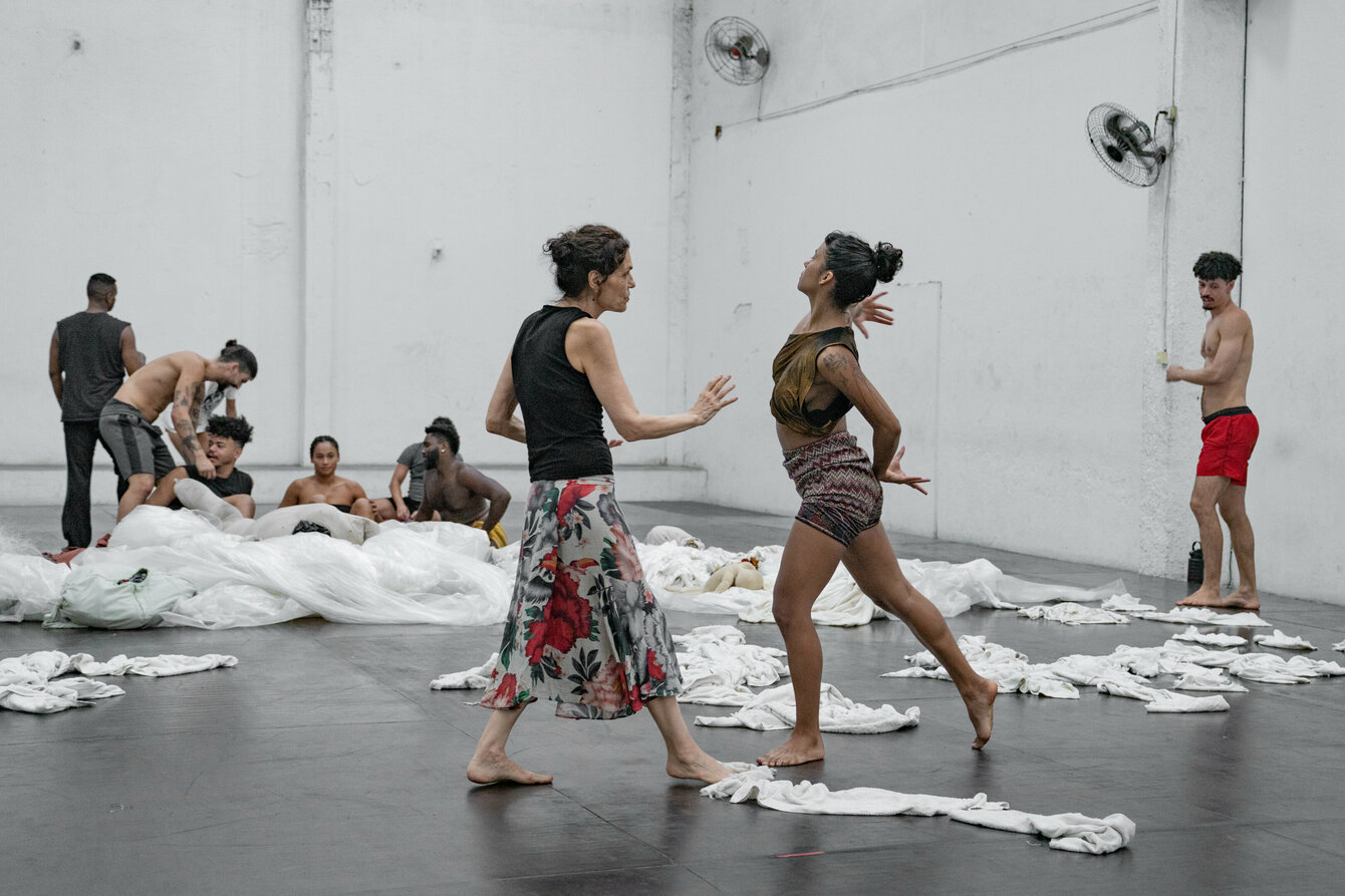 Lia Rodrigues and the performers of her piece “BORDA”. They are in a hall. In the foreground Lia Rodrigues (left), opposite to her a person performing a movement. In the background, several performers are busy with themselves. Fabrics lie on the floor.