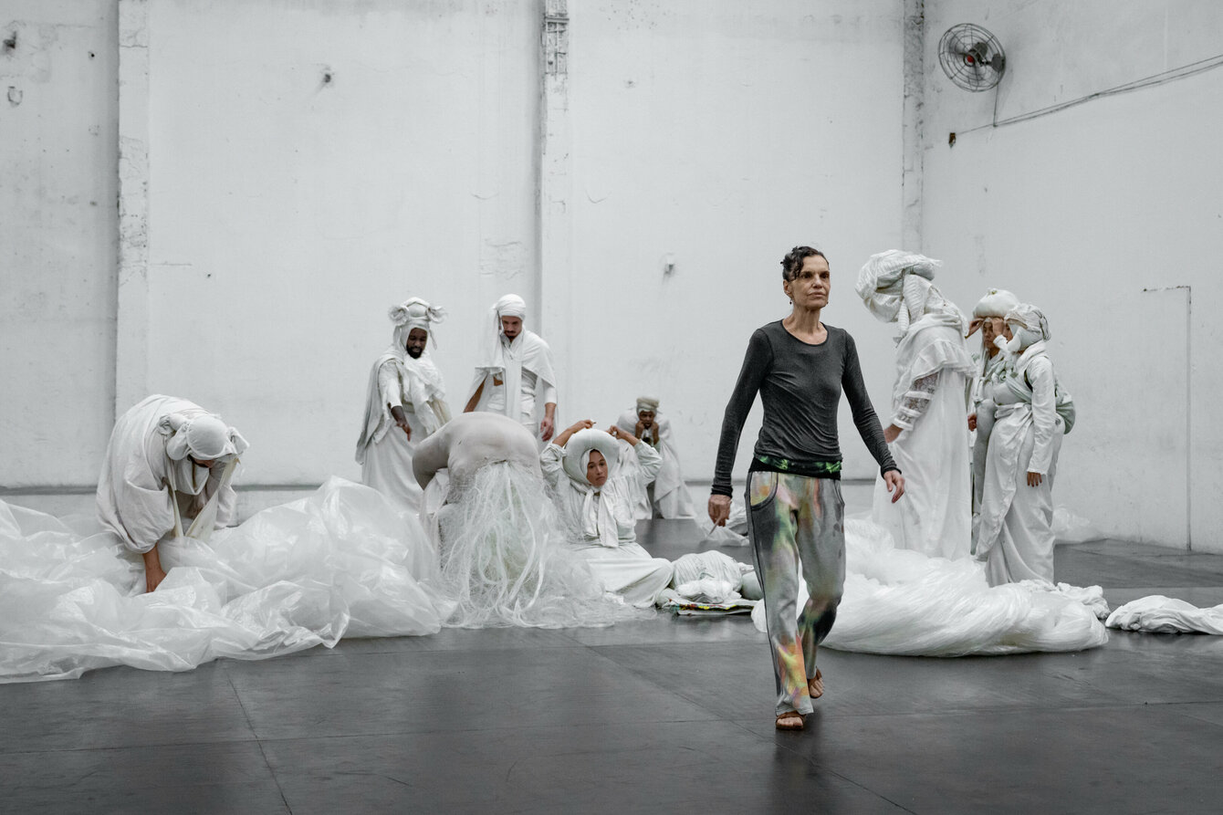 A hall, in it the choreographer Lia Rodrigues and the performers of her piece “BORDA”. Lia Rodrigues is wearing a dark top and gray trousers with a colour gradient. She walks upright to the front. Behind her are the performers, their bodies wrapped in white fabrics. They are preoccupied with themselves.  