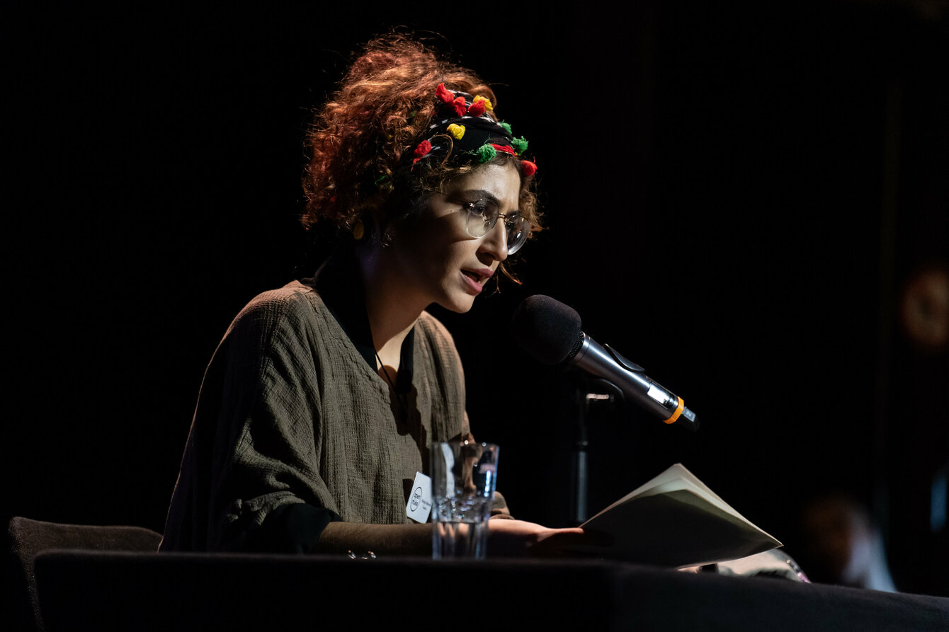 Ein Foto von Miedya Mahmod bei einer Lesung auf der Bühne. Miedya hat dunkle lockige Haare und trägt eine Brille.