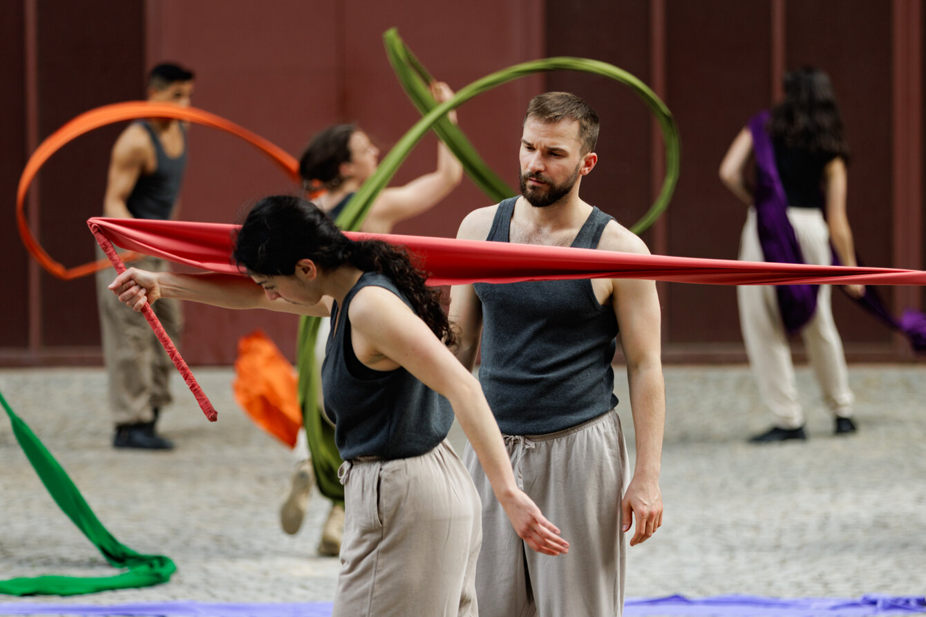 Mehrere Tänzer:innen bewegen sich in einem choreografierten Spiel mit bunten Stoffbahnen. Im Vordergrund verbindet ein rotes Tuch zwei Personen – ein Moment voller Spannung, Konzentration entsteht vor urbaner Kulisse.
