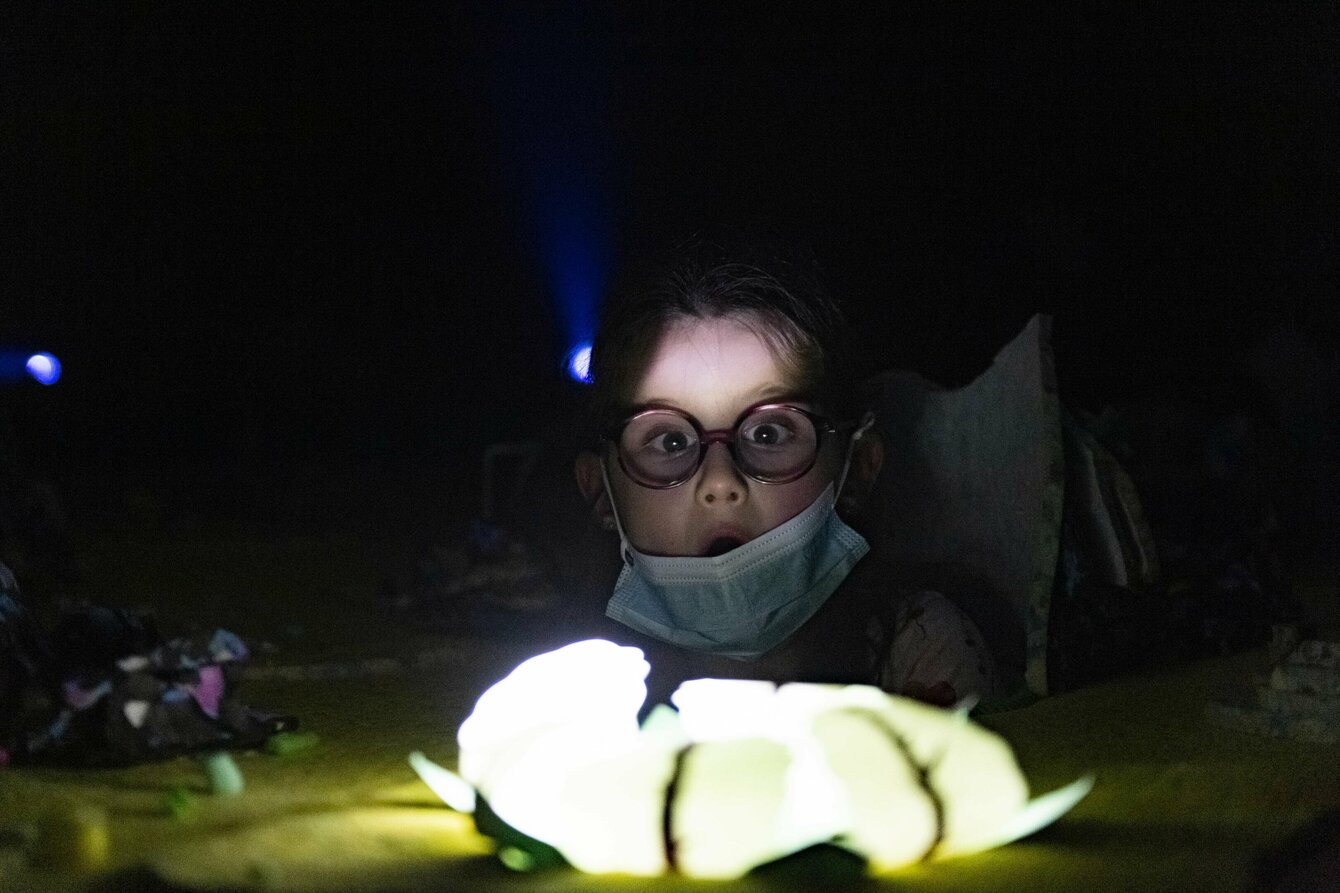 Ein Kind mit runder Brille schaut erstaunt in die Kamera, der Kopf schaut aus einem Loch in einem Bühnenbild heraus. 