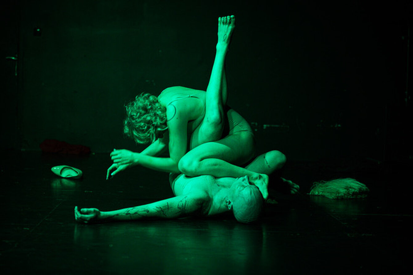 Eine Szene aus der Performance Unsex Me Here von Maria F. Scaroni. Das Bild zeigt zwei Personen, die auf dem Bühnenboden liegen und ihre Körper ineinander verschränken. Eine Person davon liegt auf dem Rücken und streckt ein Bein in die Luft. Beide Körper sind in grünes Licht getaucht. 