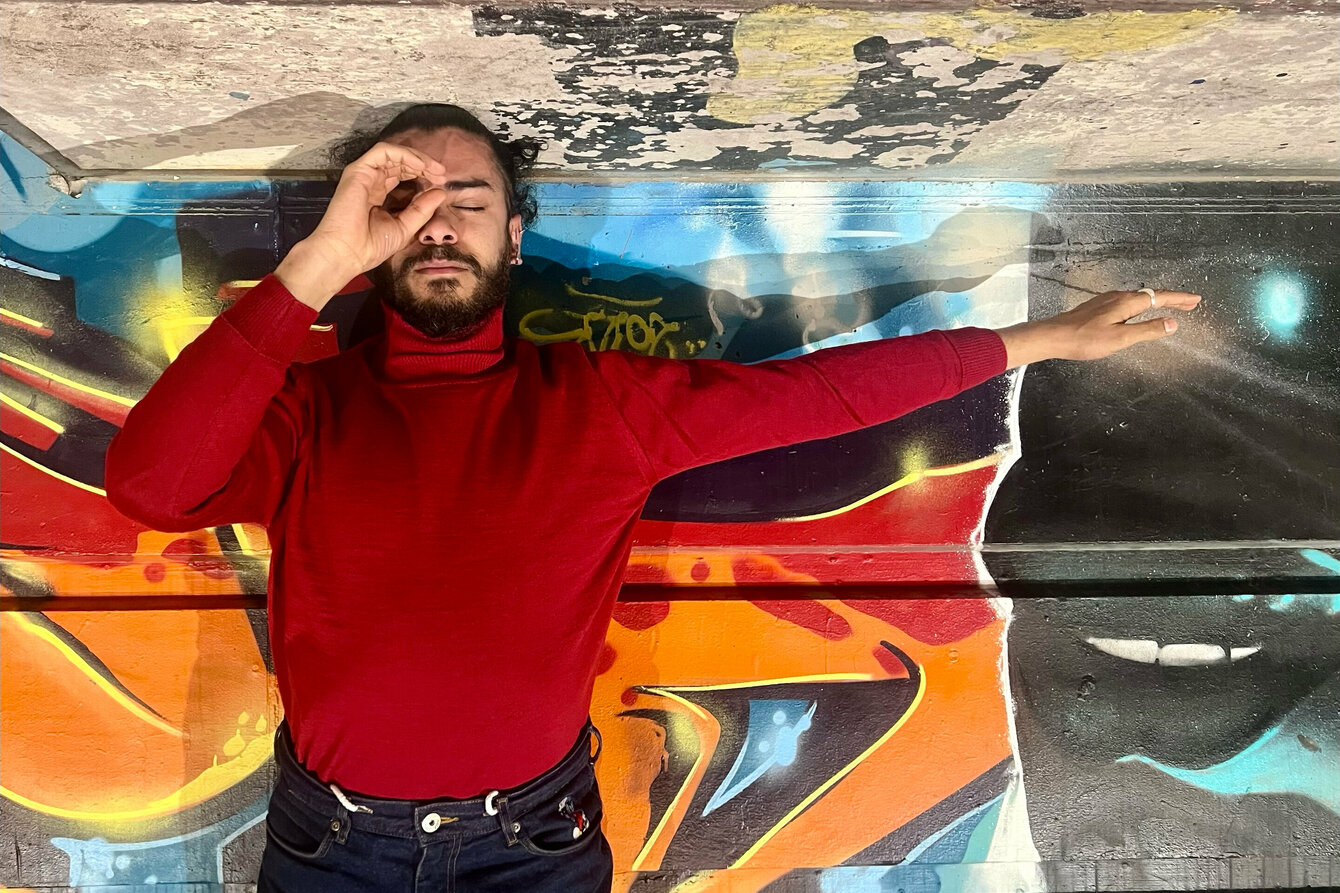 Ein Foto von Mohamed Toukabri. Er liegt auf einem Boden, der mit Graffiti gestaltet ist. Er trägt einen roten Rollkragenpullover und eine schwarze Hose und streckt seinen rechten Arm zur Seite. Er formt mit der linken Hand ein C und hält es vor das linke Auge.