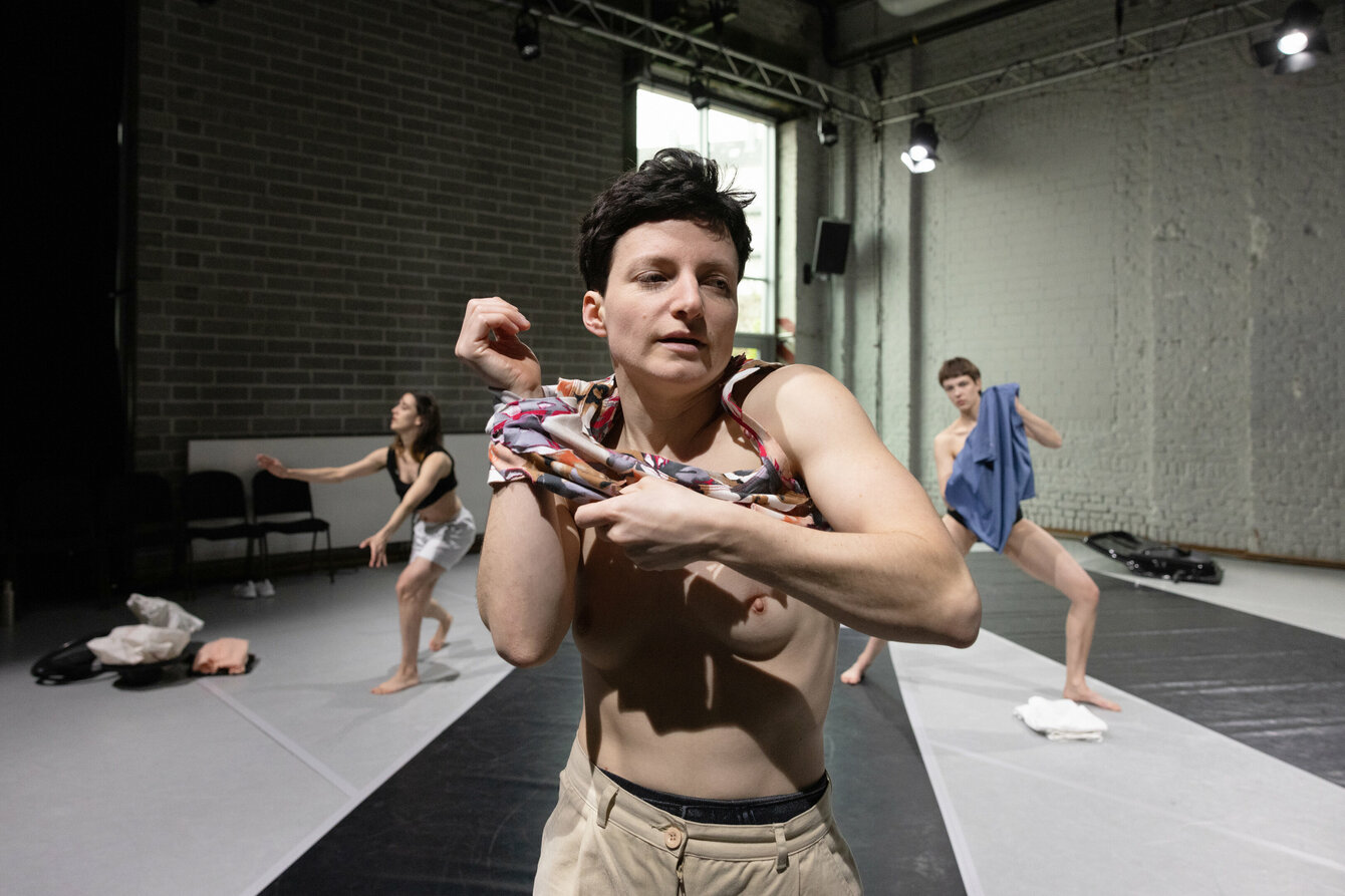 Das Bild zeigt eine weiblich gelesene Performerin, die sich in einem Tanzstudio ihr Hemd auszieht. Im Hintergrund sind zwei weitere Tänzer:innen zu sehen. Der Raum hat Backsteinwände und große Fenster. Der Tanzboden hat breite, dunkle Streifen.