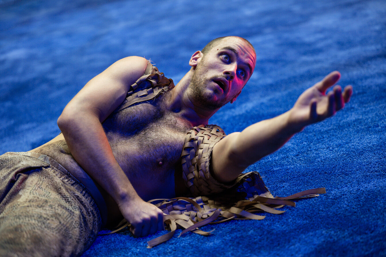 Eine Szene aus Some Thing Folk: Ein*e Performer*in liegt auf einem blauen Teppich. Die Person ist oberkörperfrei und hat eine beige Hose aus Leder an. Die Person blickt hoch und streckt dabei die Hand aus.