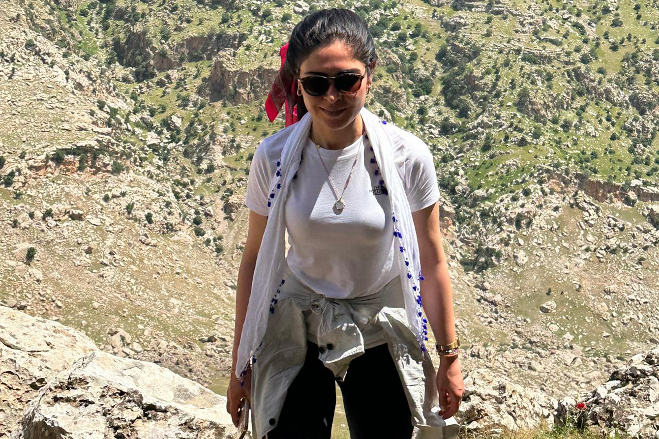 Ein Foto von Şermin Güven in einer Berglandschaft. Şermin hat dunkle Haare, die zu einem Zopf gebunden sind, ein weißes Tshirt, eine schwarze Hose sowie ein weißes Tuch um die Schultern. Sie trägt eine Sonnenbrille. 