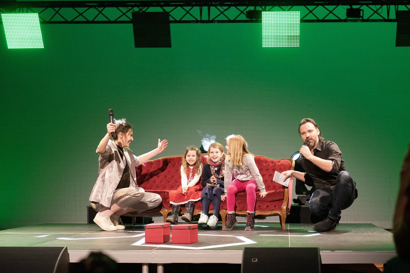 Drei Kinder sitzen auf einem roten Sofa auf der Bühne, zwei Moderator:innen sitzen außen und befragen die Kinder