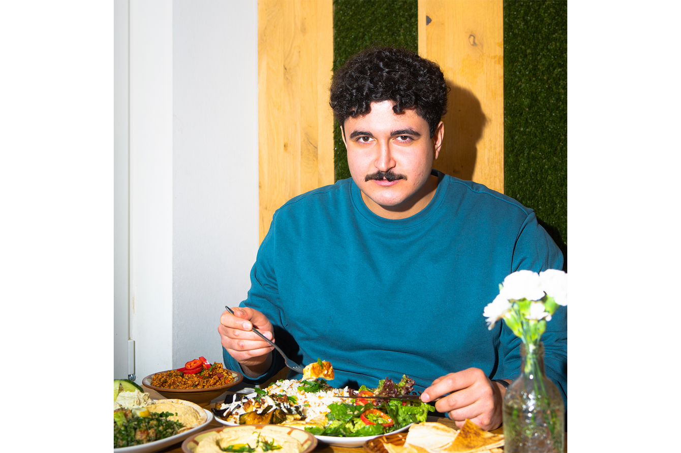 Ein Foto von Arman Marvani. Arman hat kurze dunkle, lockige Haare und einen Schnurrbart. Er trägt einen blauen Pullover und sitzt an einem gedeckten Tisch. In der Hand hält er eine Gabel und blickt in die Kamera. Vor ihm auf dem Tisch sind mehrere Teller mit Speisen zu sehen. 