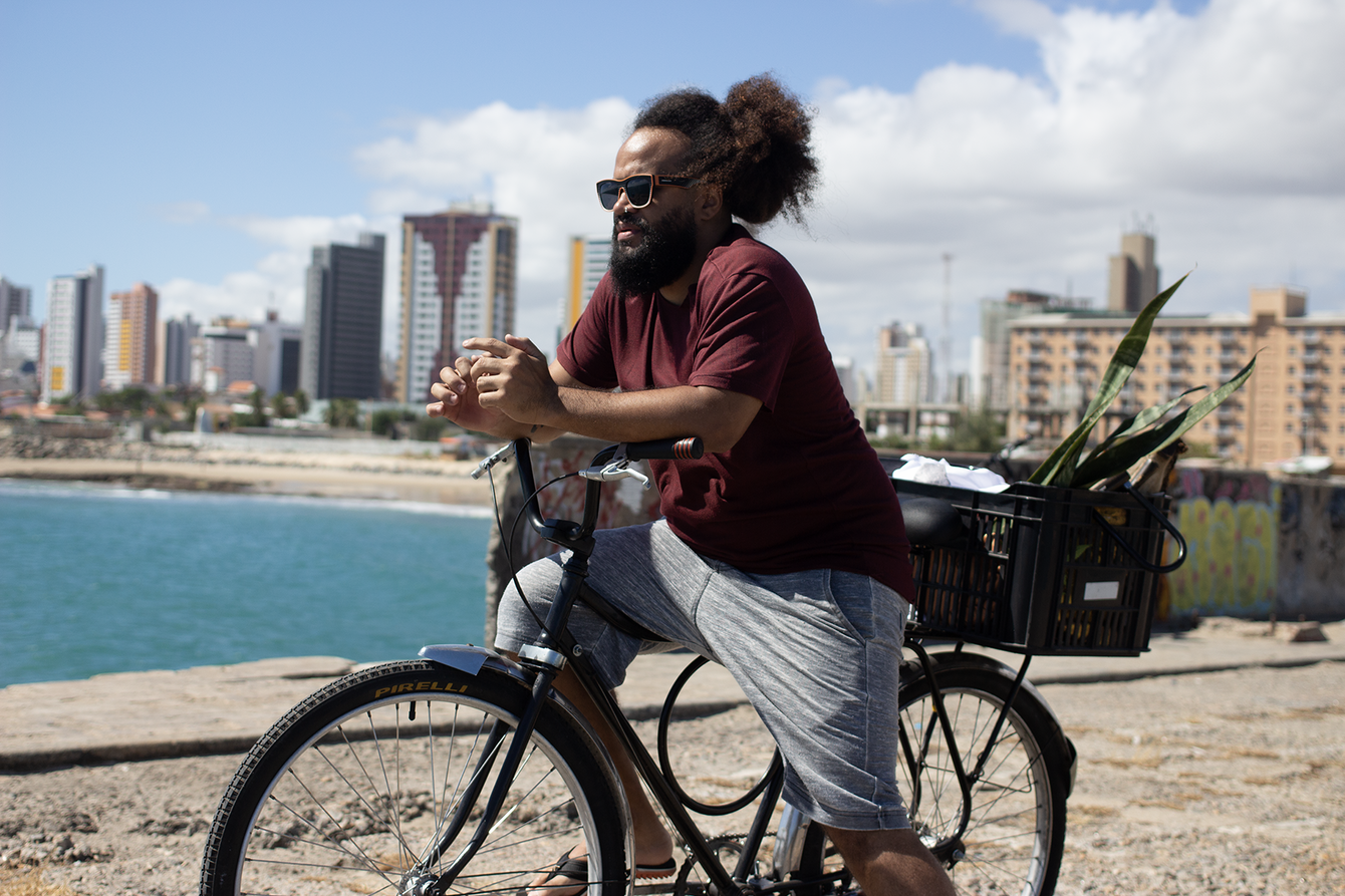 Ein Mann steht mit einem Fahrrad an einer Uferpromenade. Im Hintergrund sieht man Wasser, Strand und Hochhäuser.