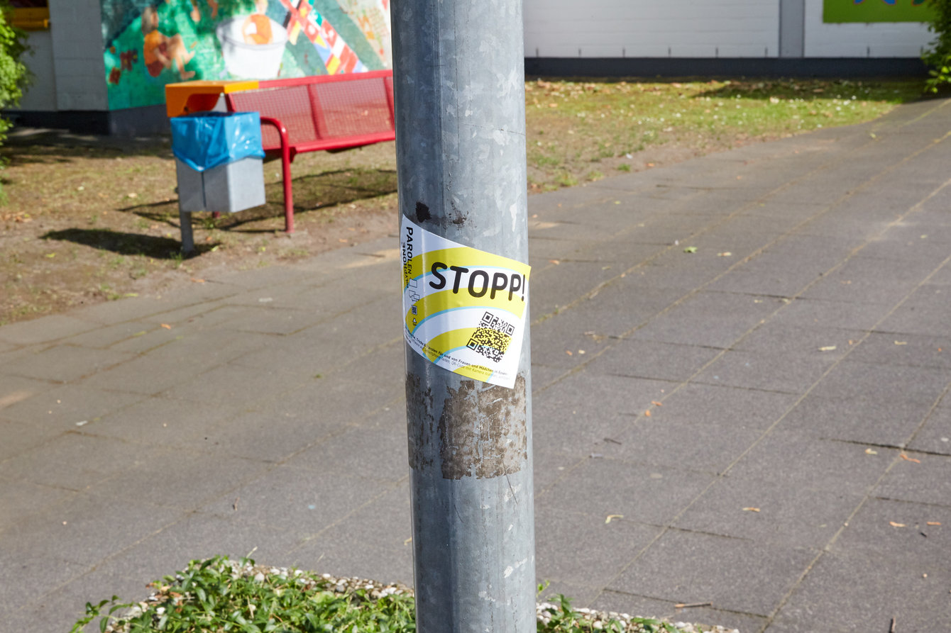 Eine Laternenpfahl mit Aufkleber ist mittig auf dem Bild im Vordergrund, im Hintergrund eine rote Parkbank mit Mülleimer 