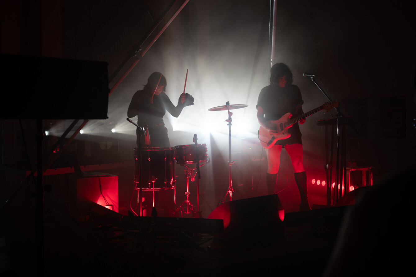 Zwei Musikerinnen stehen auf einer Bühne und sind in rotes Licht gehüllt. Ihre Silhouetten werden von einem Scheinwerfer hinter der Bühne angestrahlt.