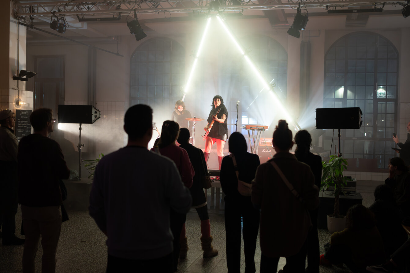 In einem großen, offenen Raum spielt eine Band auf einer Bühne. Die Musikerinnen sind in Nebel gehüllt und werden von Leuchtröhren hinter der Bühne angestrahlt. Vor der Bühne steht das Publikum im Raum verteilt.