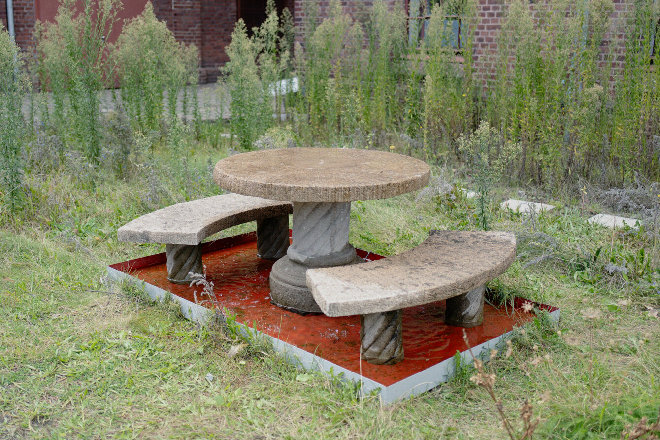 Eine Brunneninstallation auf der begrünten Fläche vor PACT Zollverein. Es handelt sich um einen runden Tisch aus Stein, aus dessen Mitte Wasser sprudelt. Drumherum zwei Bänke. Tisch und Bänke stehen in einem roten Becken.