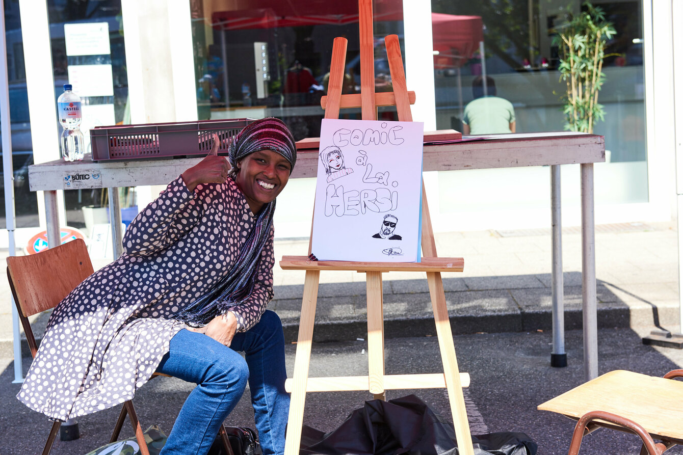 Eine Person die neben einer gezeichneten Karikatur sitzt und lächelt und den Daumen hebt