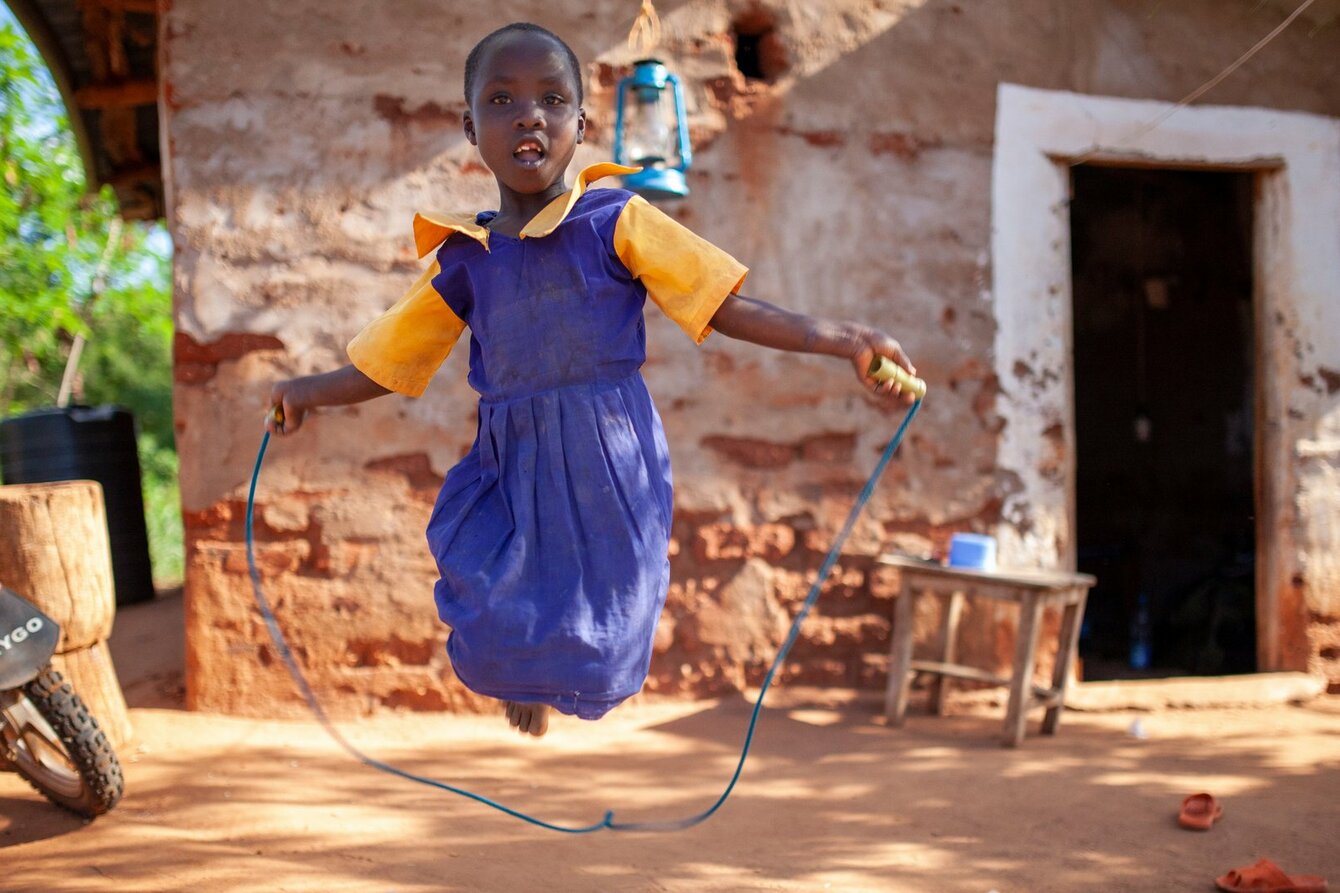 Ein Still aus dem Film "With Grace": Es zeigt ein Kind beim Seilspringen, draußen vor einem Haus. Das Kind trägt über einem gelben Tshirt ein blaues Kleid. 