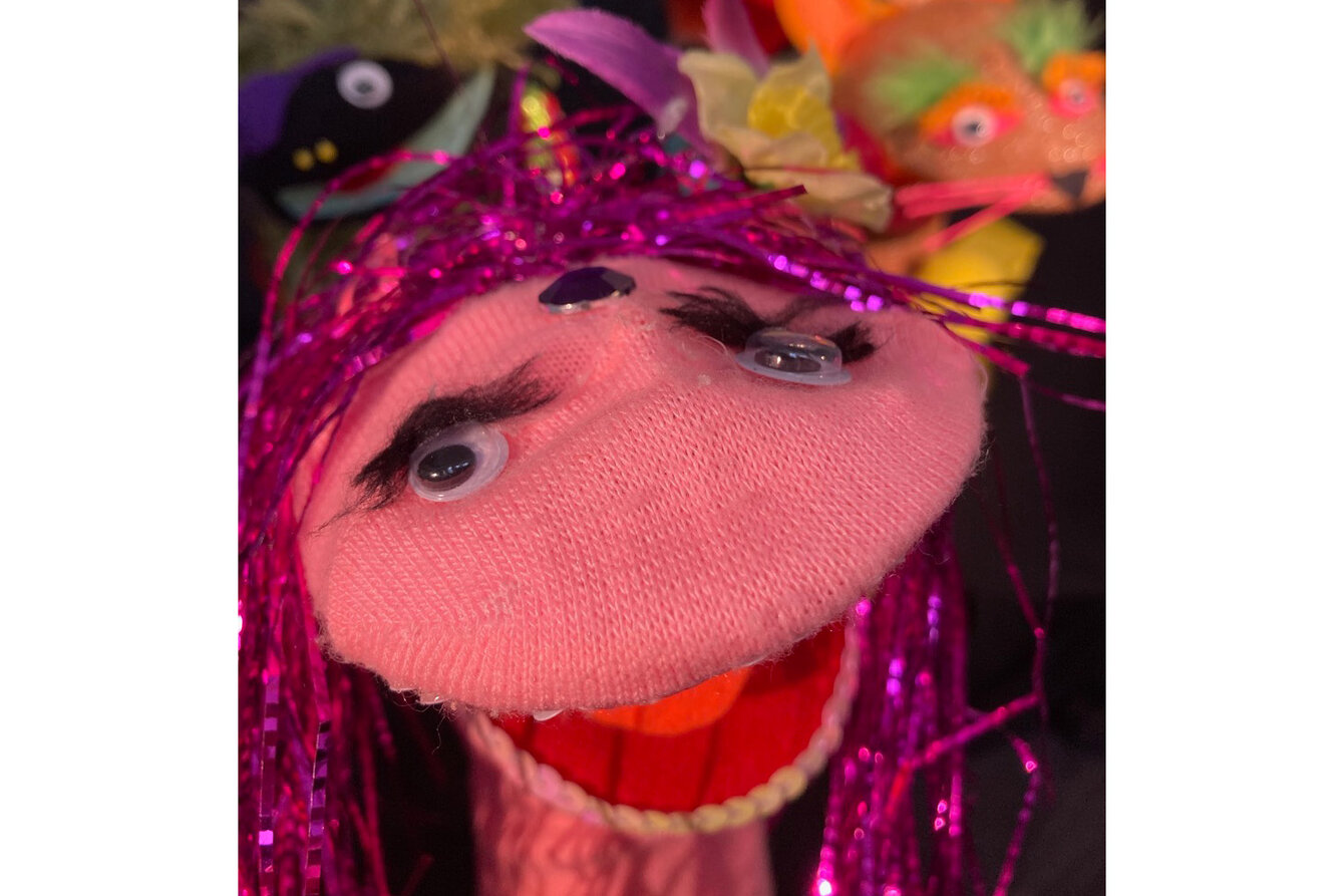 Ein Foto von bunten Puppen, die aus Socken erstellt wurden. Im Vordergrund eine rosafarbene Sockenpuppe mit pinken Lametta-Haar, Augen und Mund. Dahinter weitere bunte Exemplare.