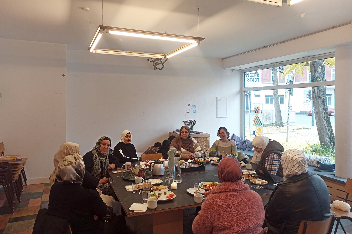 Eine Gruppe Frauen sitzt in der WerkStadt um einen Tisch herum. Die meisten Personen tragen ein Kopftuch. Der Tisch ist zu gedeckt, Speisen und Getränke stehen bereit. Im Hintergrund spielen Kinder. 