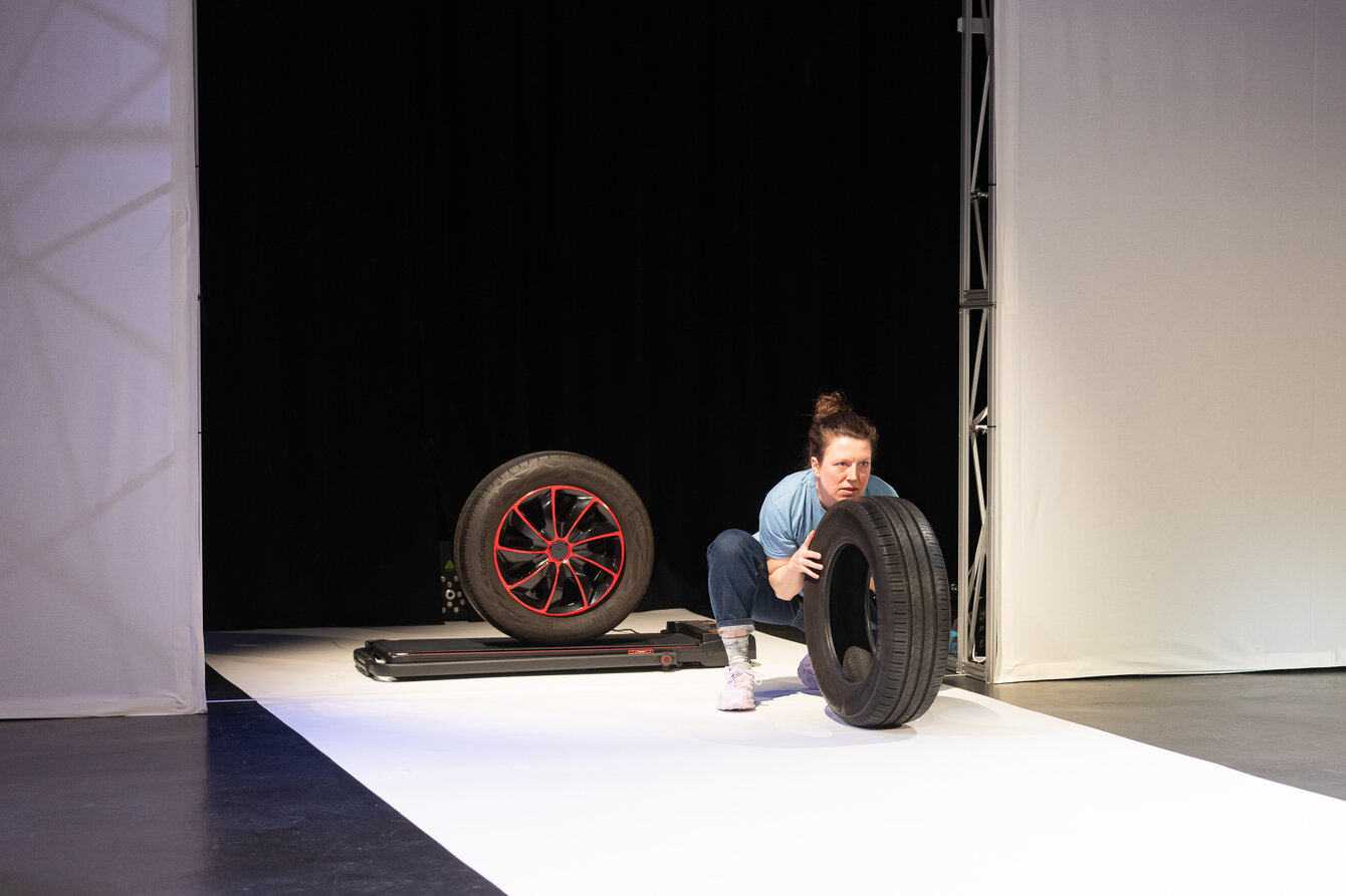 Auf einer Bühne rollt ein ein Autoreifen auf einem elektrischen Laufband. Davor hockt eine Performerin Verena Brakonier mit einem weiteren Reifen.